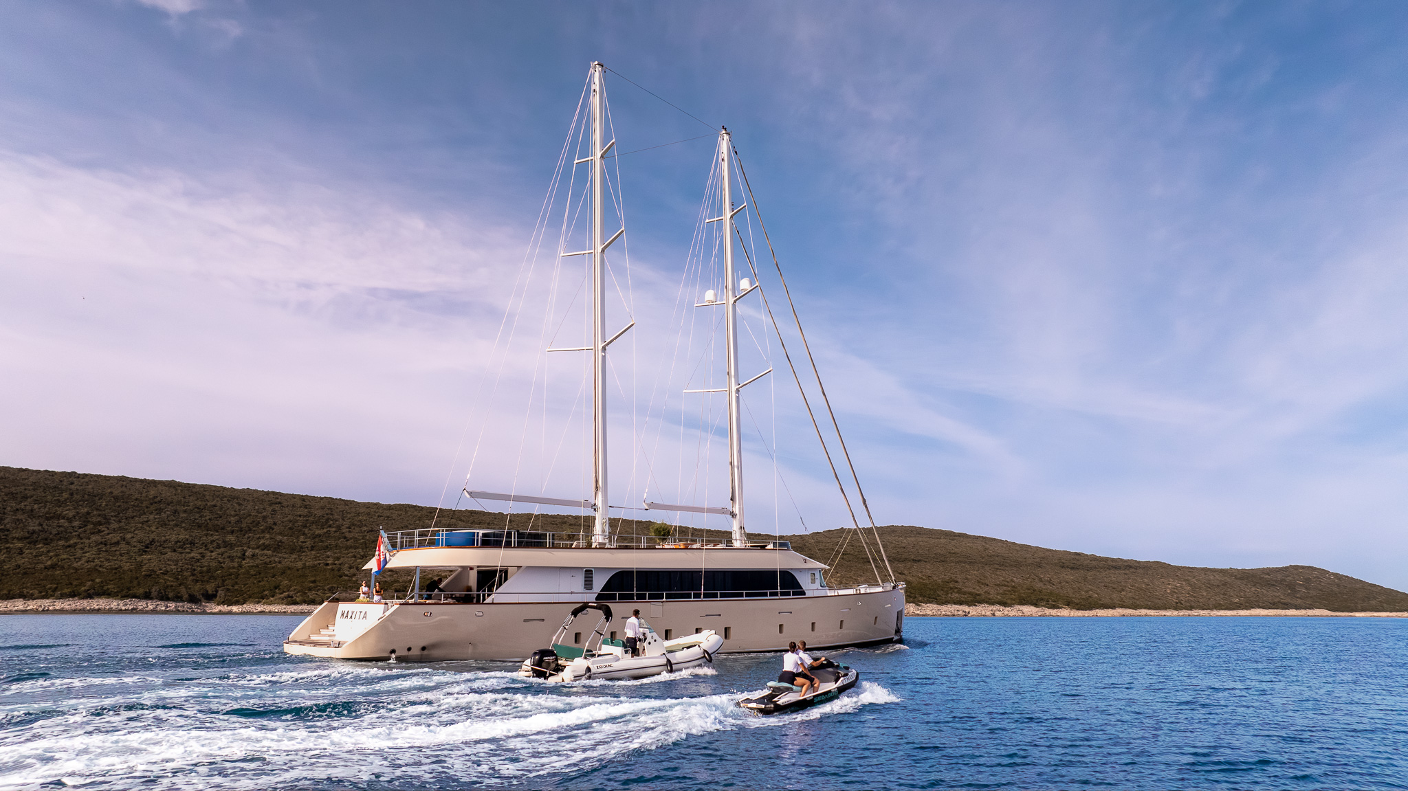 MAXITA A large, modern catamaran yacht with two tall masts sails near a coastline, accompanied by a small inflatable boat and a jet ski on clear blue water under a partly cloudy sky.