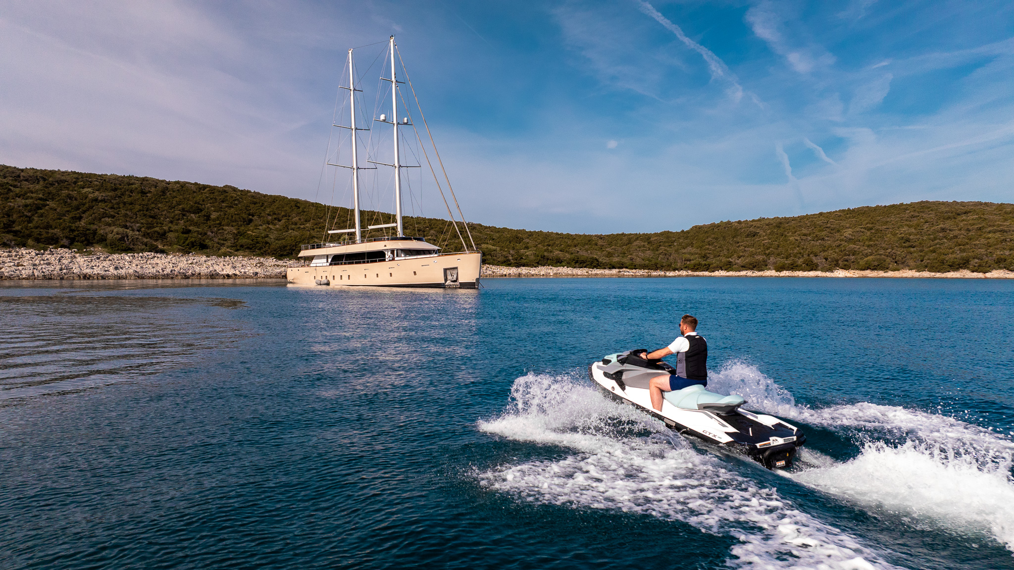 MAXITA A person rides a jet ski on calm blue water toward a large luxury yacht anchored near a green, tree-covered shoreline under a partly cloudy sky.