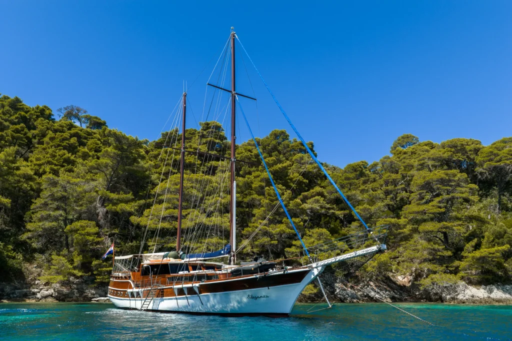 ELEGANZA A white sailboat with wooden trim is anchored in clear blue water near a lush, green, tree-covered shoreline under a bright blue sky.