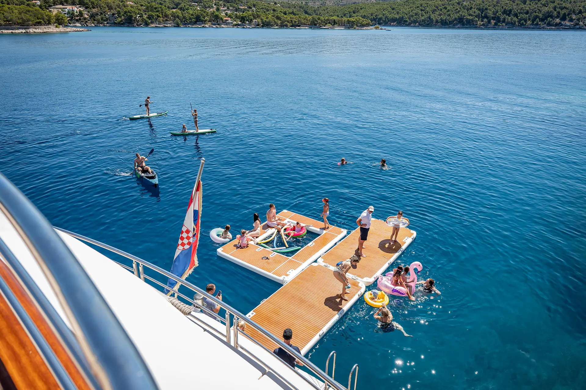 People relax and play on floating docks and paddleboards in clear blue water near a yacht, with swimmers and inflatables; a Croatian flag is visible.