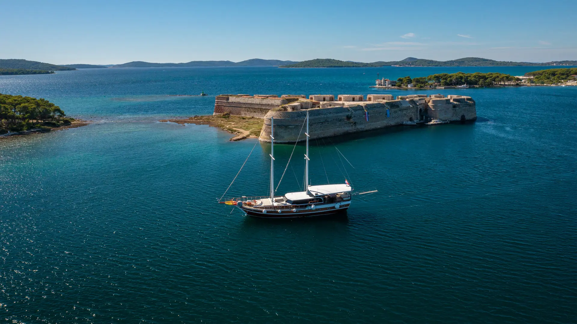A sailboat floats near a historic stone fortress on a small island, surrounded by blue water and distant hills under a clear sky.