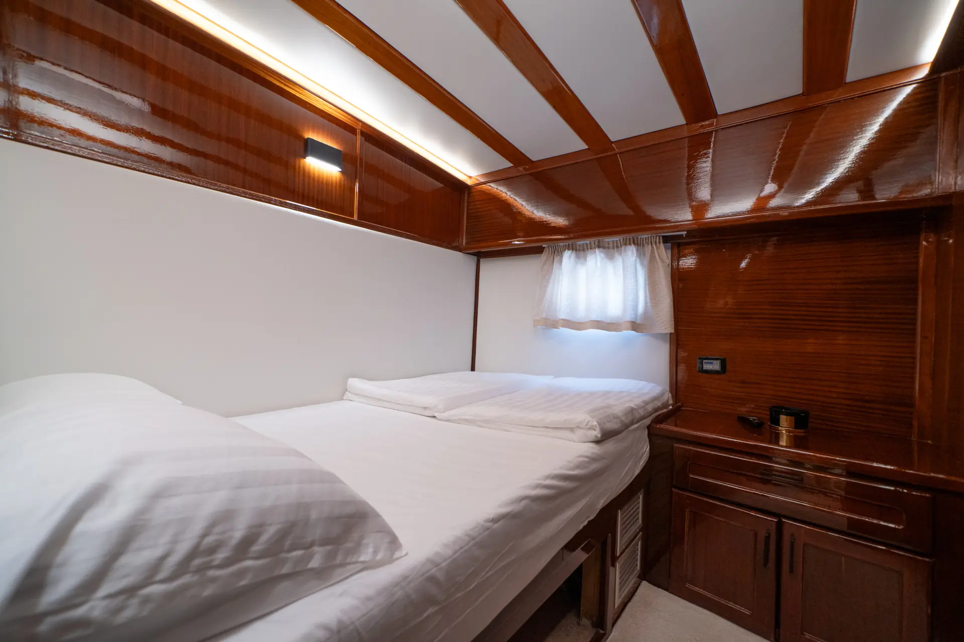 A small, cozy room with a single bed, white bedding, wooden walls and ceiling, a small window with a sheer curtain, and built-in cabinets.