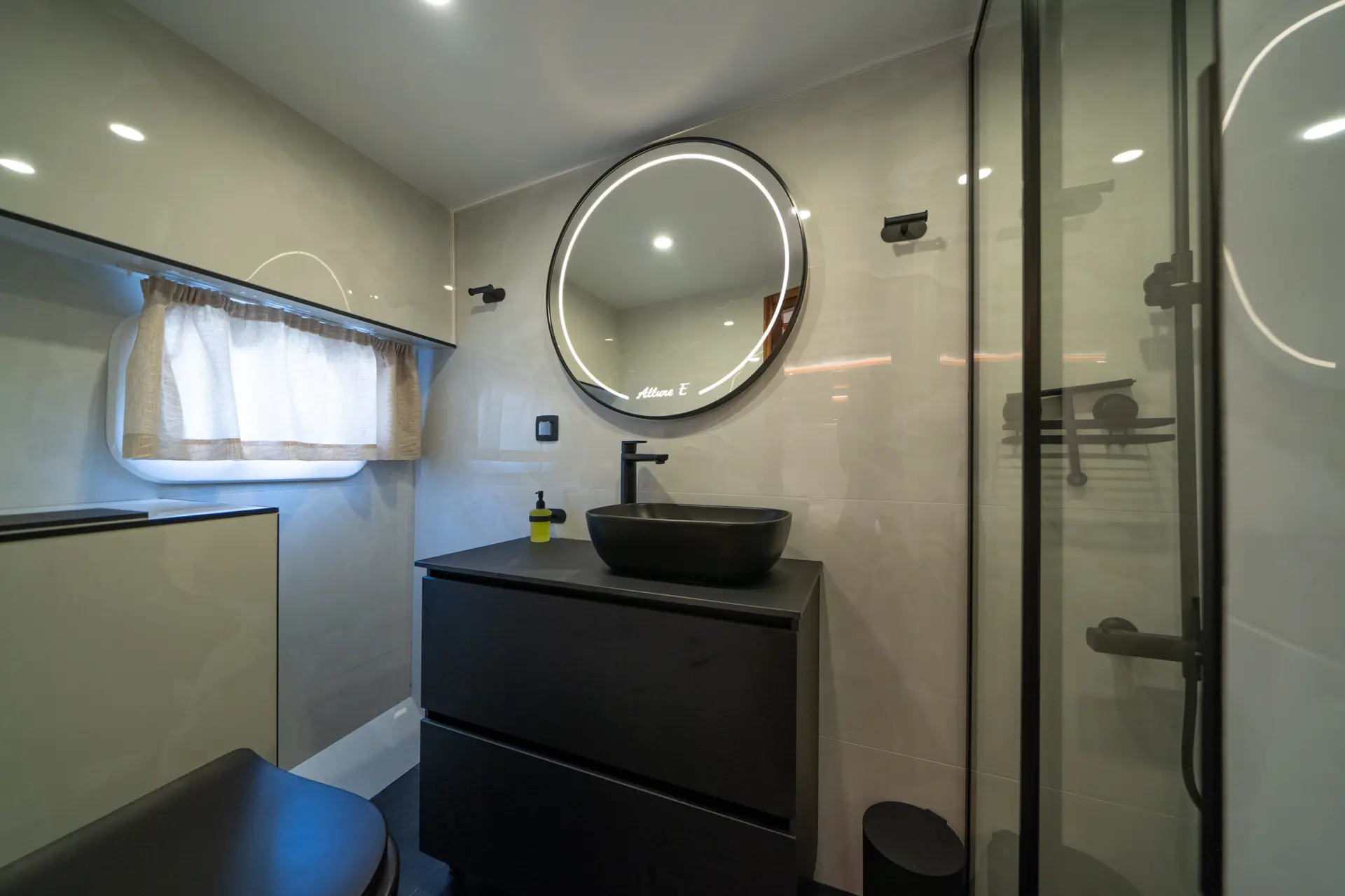 Modern bathroom with circular backlit mirror, black sink and faucet on vanity, glass shower door, and a small window with a light curtain. Minimalist and sleek design in neutral tones.