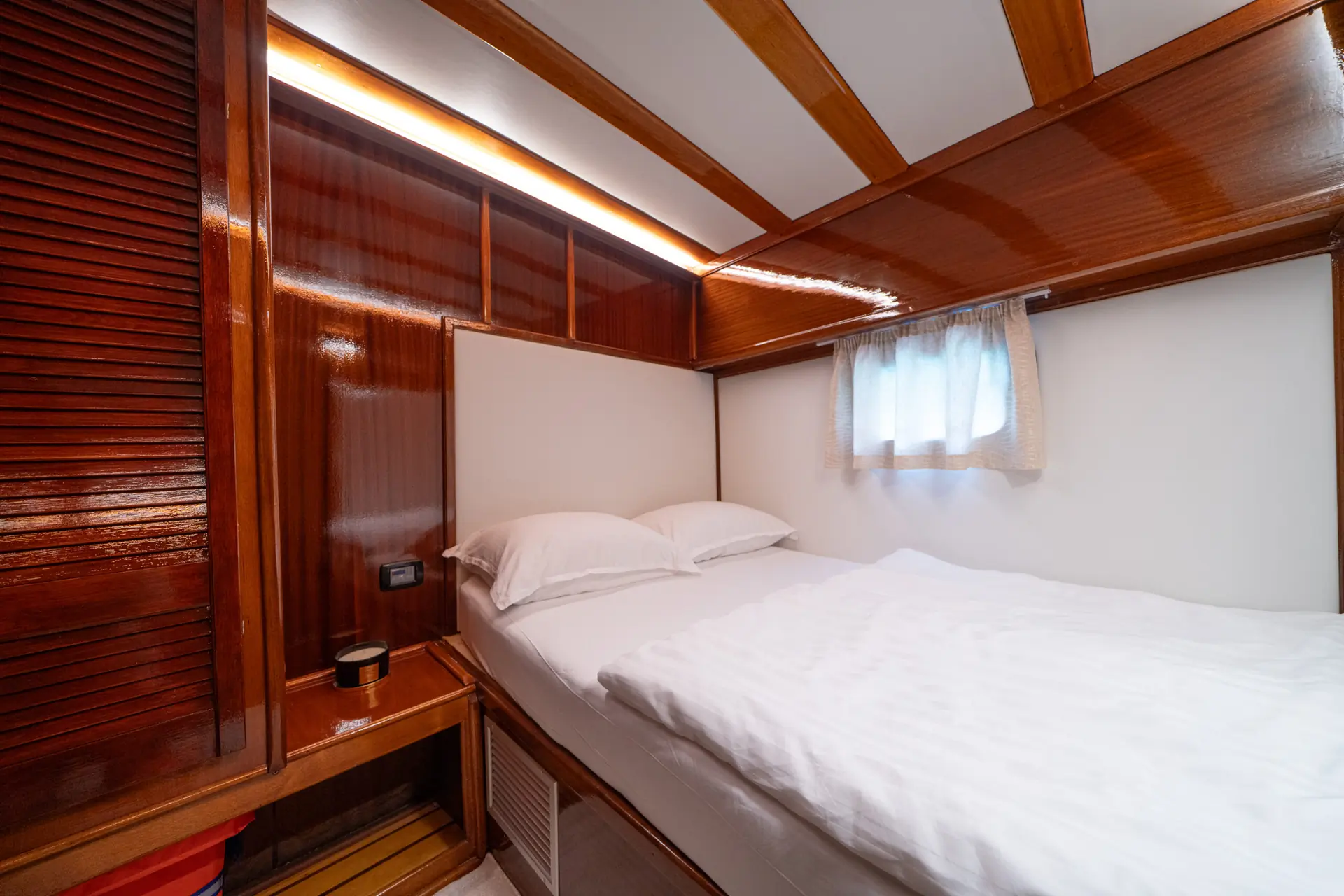 Cozy cabin bedroom with wood paneling, a double bed with white bedding, a small curtained window, and warm ambient lighting.