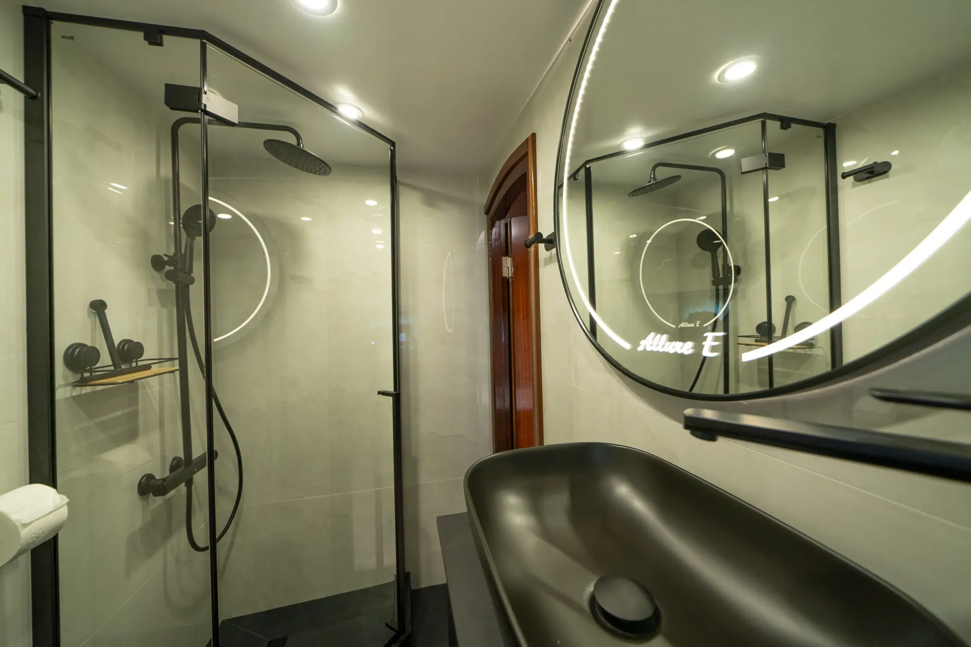 Modern bathroom with a glass shower enclosure, black fixtures, a round lighted mirror, a black sink, and white tile walls. The space appears clean and stylish with contemporary design elements.