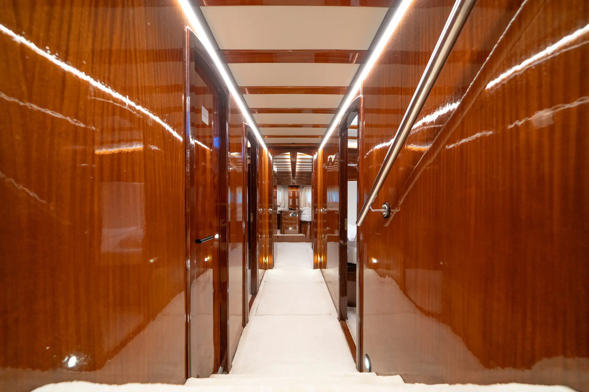 A hallway with glossy wooden walls, several doors on each side, a white carpeted floor, and ceiling lights creating a modern, luxurious interior design.
