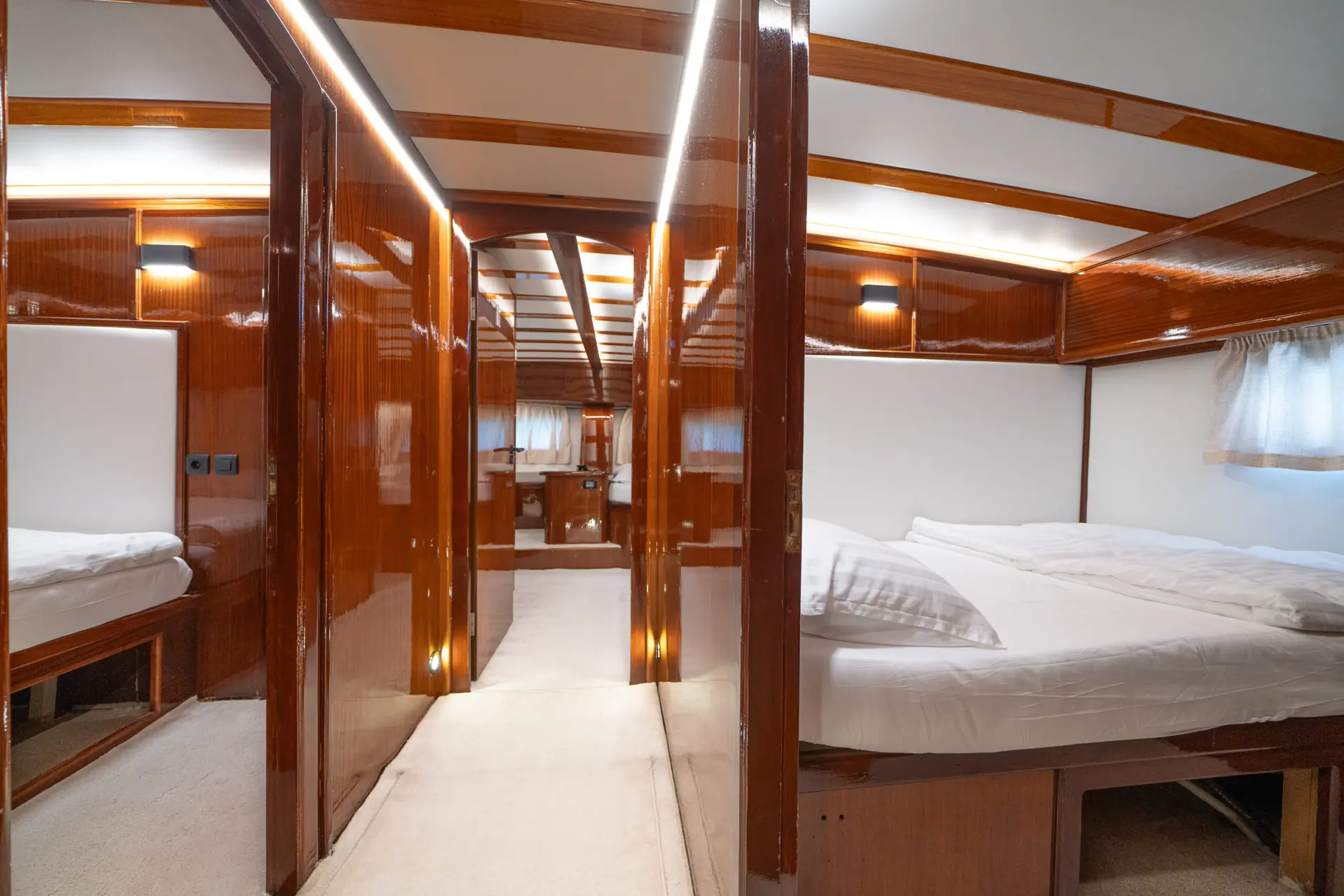 A polished wooden yacht cabin hallway with two bedrooms on each side, each with a neatly made bed, white linens, and small windows. The corridor leads to another room at the end, brightly lit.