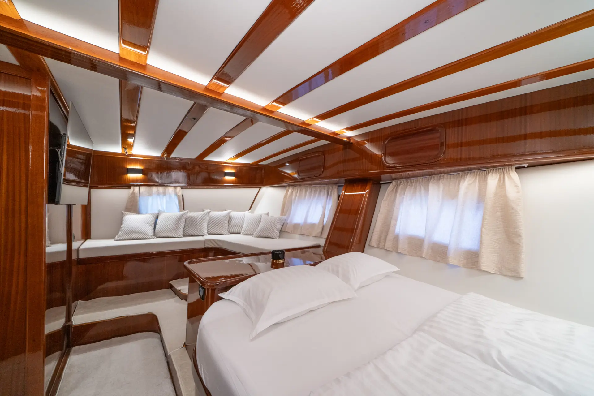 Luxurious yacht cabin with polished wood walls and ceiling, a large bed with white linens, windows with cream curtains, and a corner seating area filled with white pillows. Warm lighting creates a cozy, elegant atmosphere.