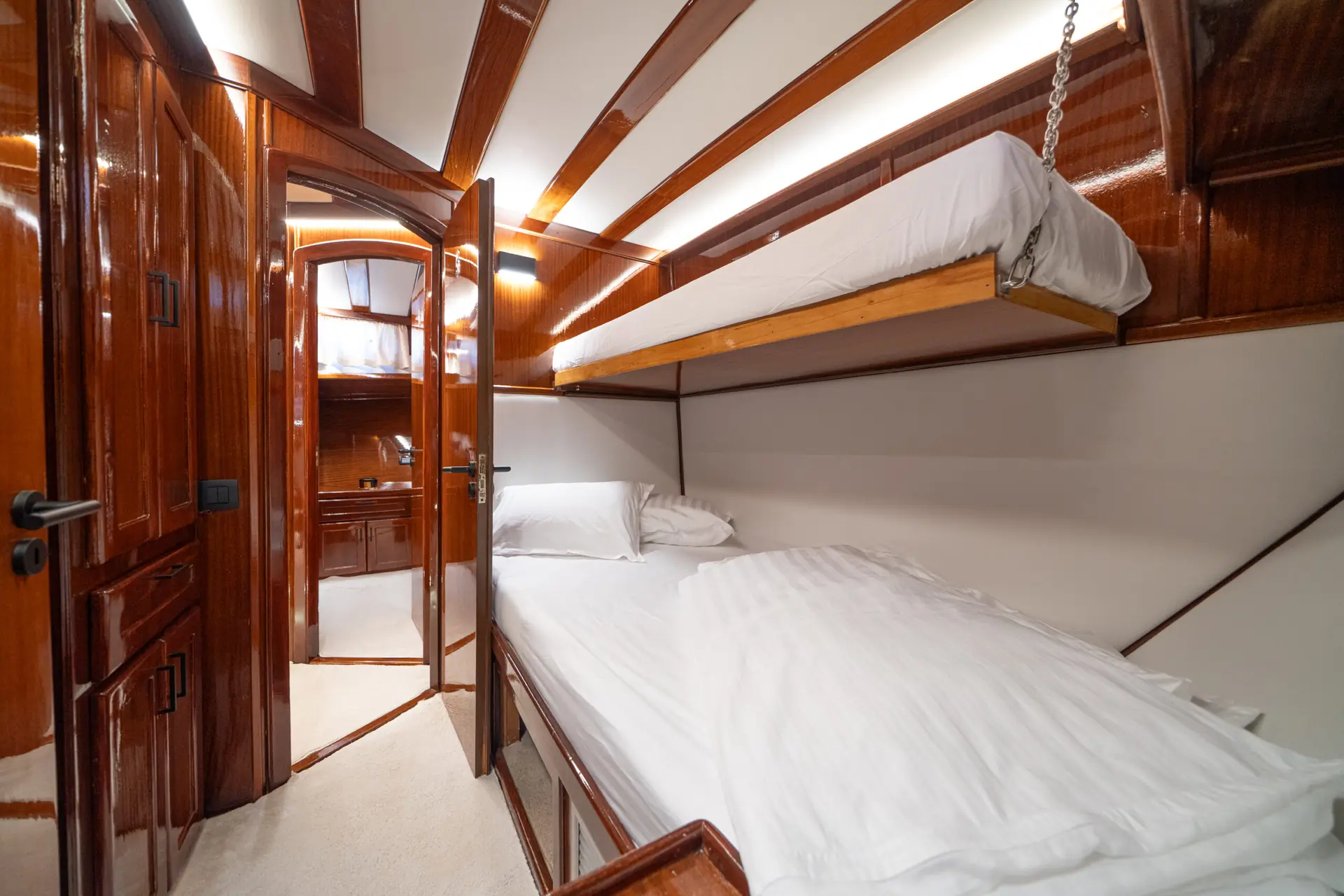 A yacht cabin with glossy wooden walls, a lower single bed made with white linens, and an upper bunk. The door is open, revealing more wood surfaces and a mirror in the adjoining room.