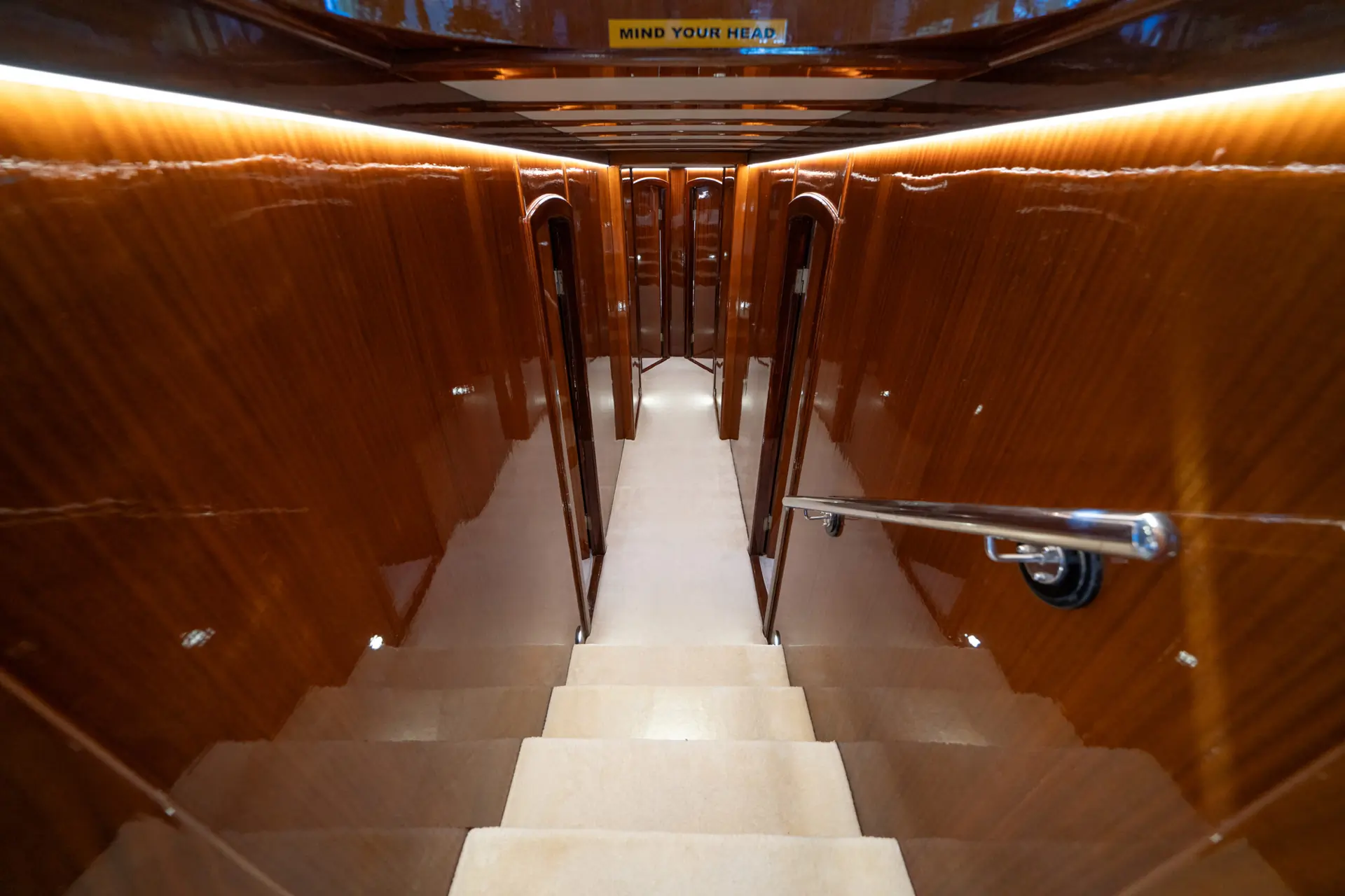Polished wood-paneled stairway with cream steps descends to a hallway. A MIND YOUR HEAD sign is visible above the corridor entrance at the bottom. Handrails line the walls, and doors are on either side of the hallway.