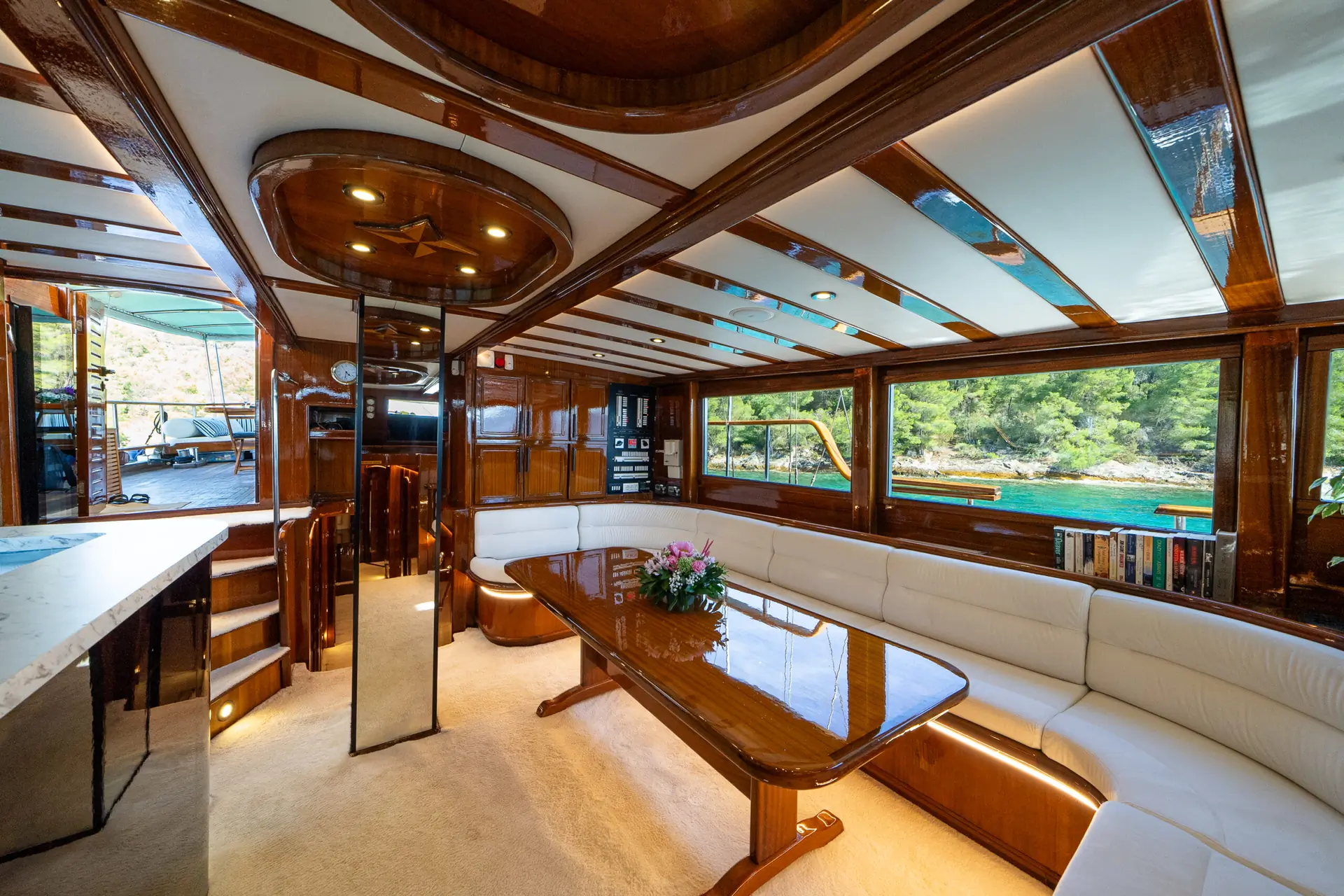 Luxurious yacht interior with polished wood, cream sectional sofa, a glossy dining table with flowers, large windows showing water and trees, and stairs leading to another deck.