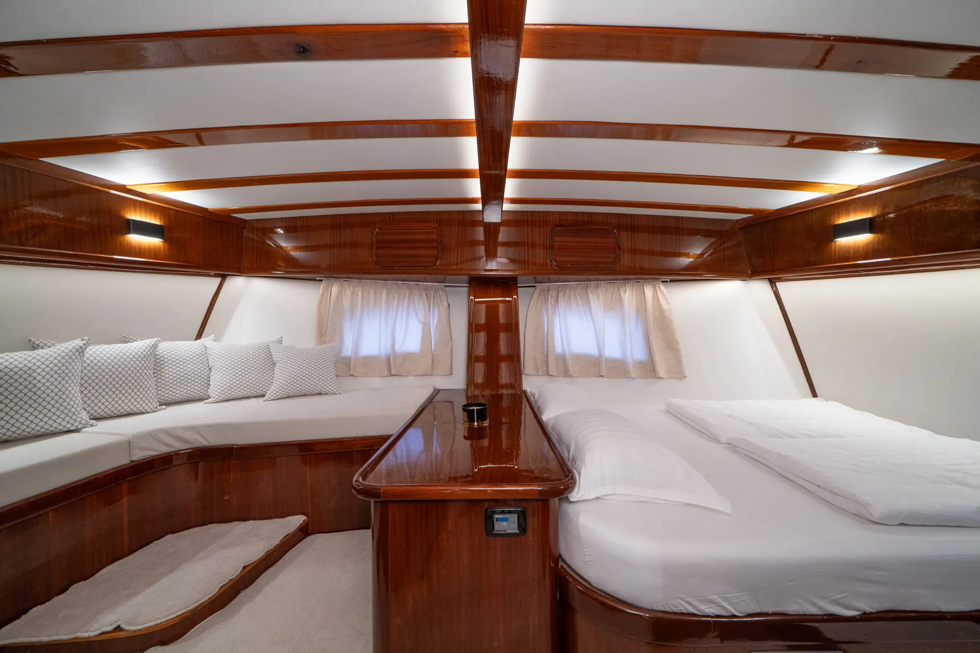 A cozy yacht cabin with polished wood, a double bed on the right, a cushioned seating area with pillows on the left, white linens, soft curtains, and ambient wall lights, creating a warm and inviting atmosphere.