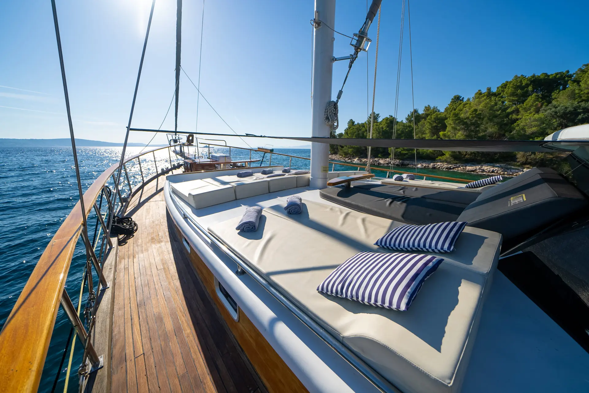 A yacht deck with sun loungers, striped pillows, and rolled towels overlooks calm blue sea and a green, tree-lined shore under a clear, sunny sky.