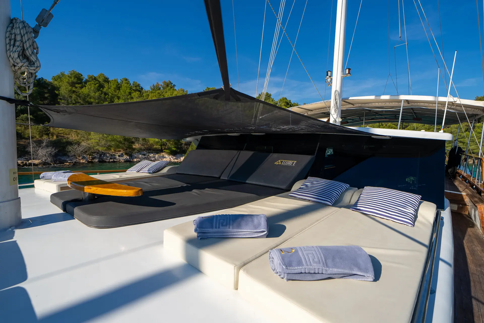 Two sun loungers and cushioned seating with striped pillows and rolled towels on a yacht deck, shaded by a black canopy, with trees and blue sky in the background.