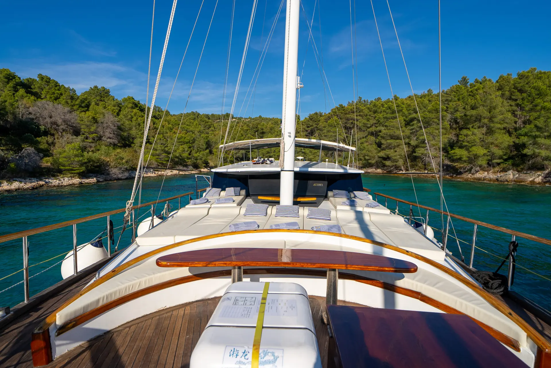 The view from a yacht deck shows sun loungers, blue waters, and a forested shoreline under a clear blue sky.