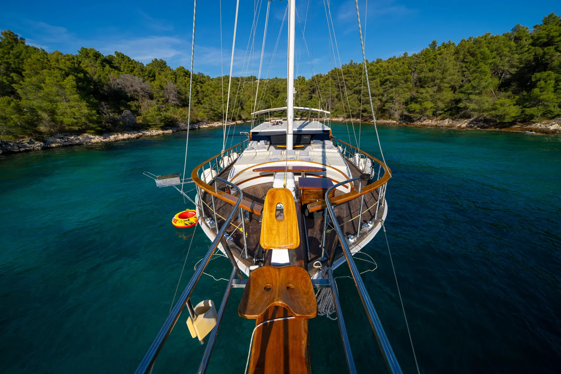 View from the bow of a wooden sailboat anchored in clear blue water, surrounded by green forested shoreline under a clear blue sky.