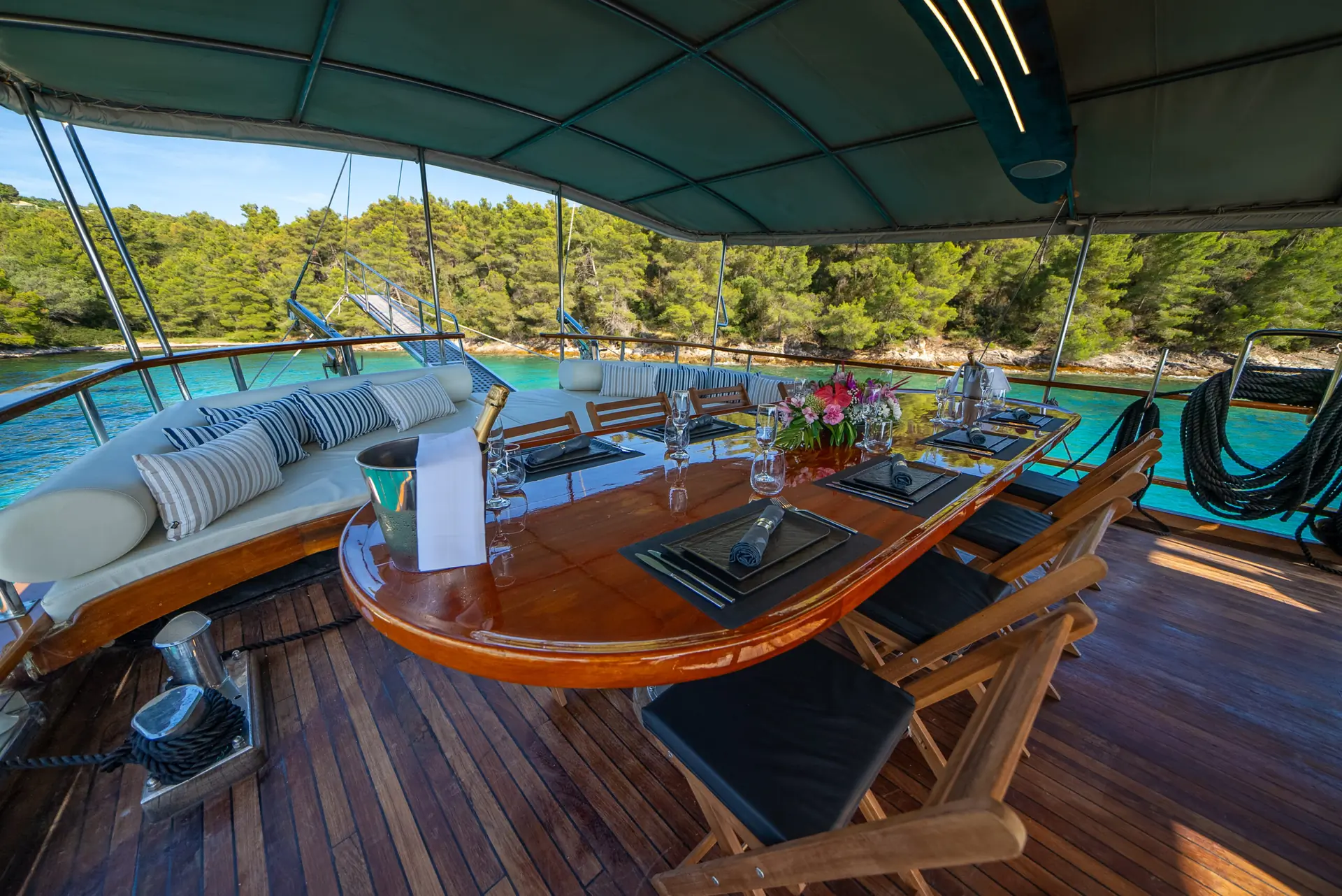 A wooden yacht deck with a dining table set for six, surrounded by chairs and cushioned benches, overlooks turquoise water and pine trees under a shaded canopy.