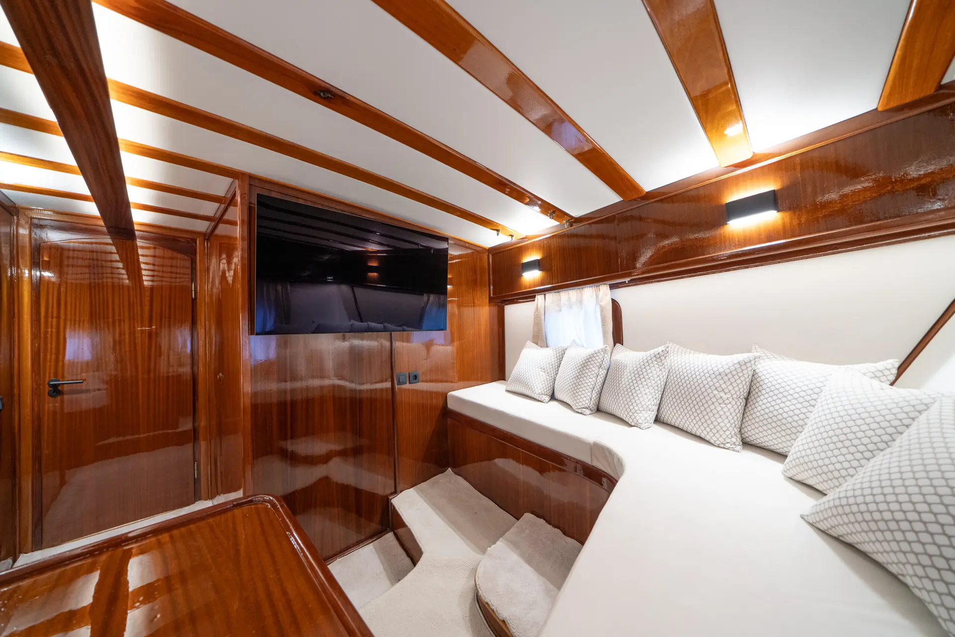 Luxurious yacht cabin with polished wood walls, a large flat-screen TV, white cushioned seating, patterned pillows, and soft lighting creating a cozy, elegant atmosphere.