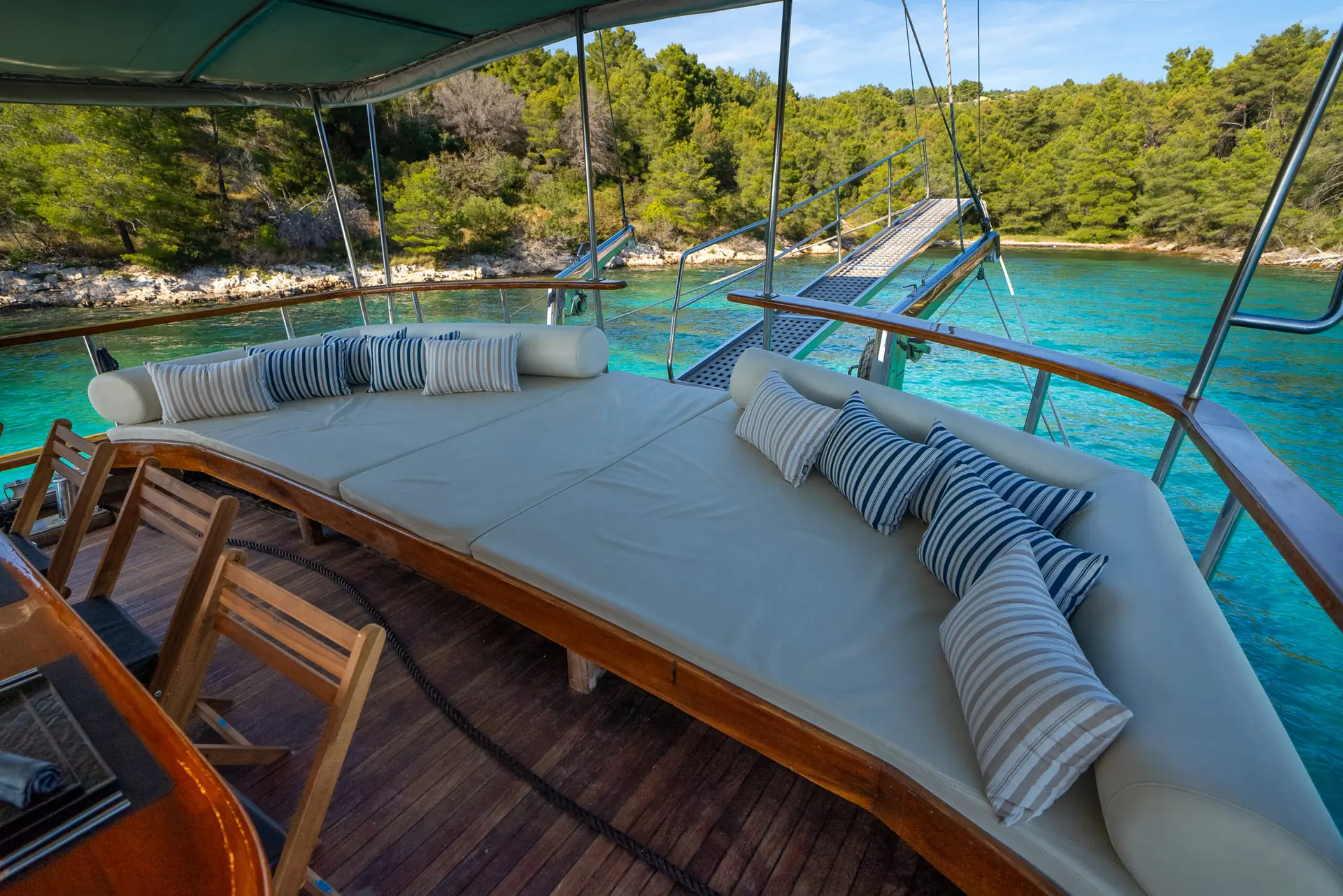 Large cushioned lounging area with striped pillows on a boat deck, overlooking clear turquoise water and a forested shoreline under a sunny sky.