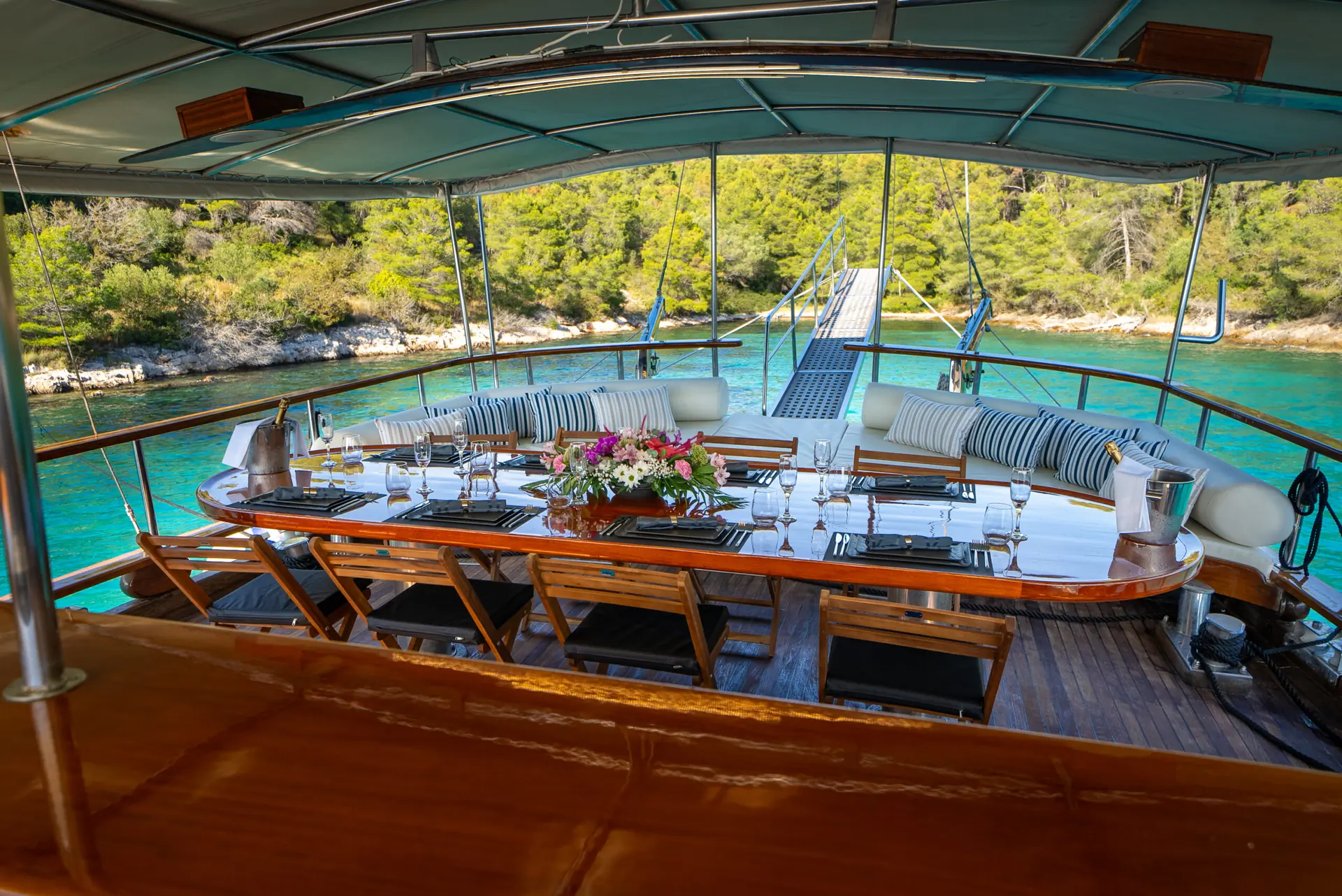 A luxurious outdoor dining area on a yacht features a long table set for a meal, wooden chairs, cushioned benches, and overlooks turquoise water and lush green trees in the background.