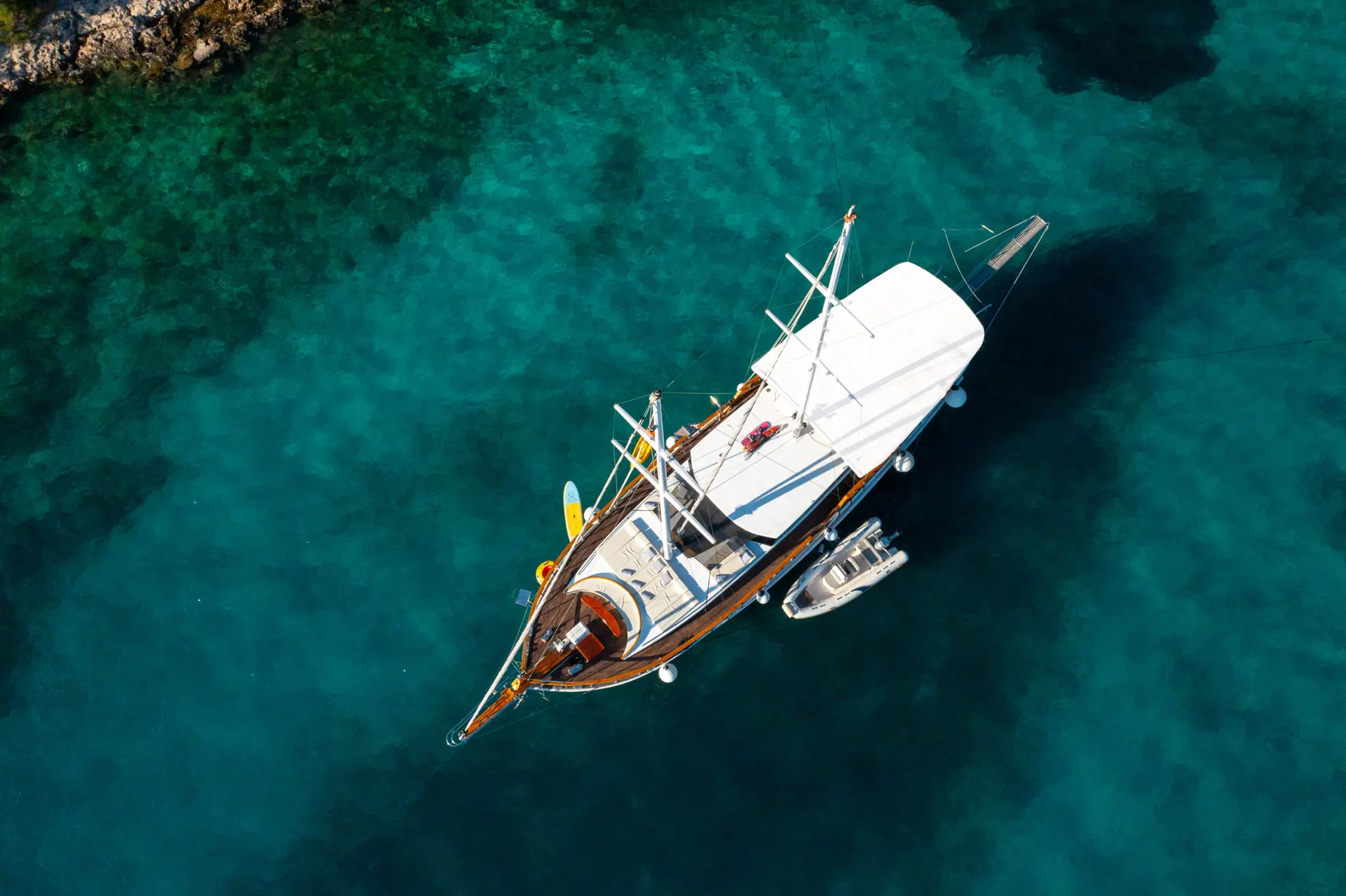 Aerial view of a white sailboat anchored in clear turquoise water near a rocky shoreline, with a small inflatable boat tied alongside.