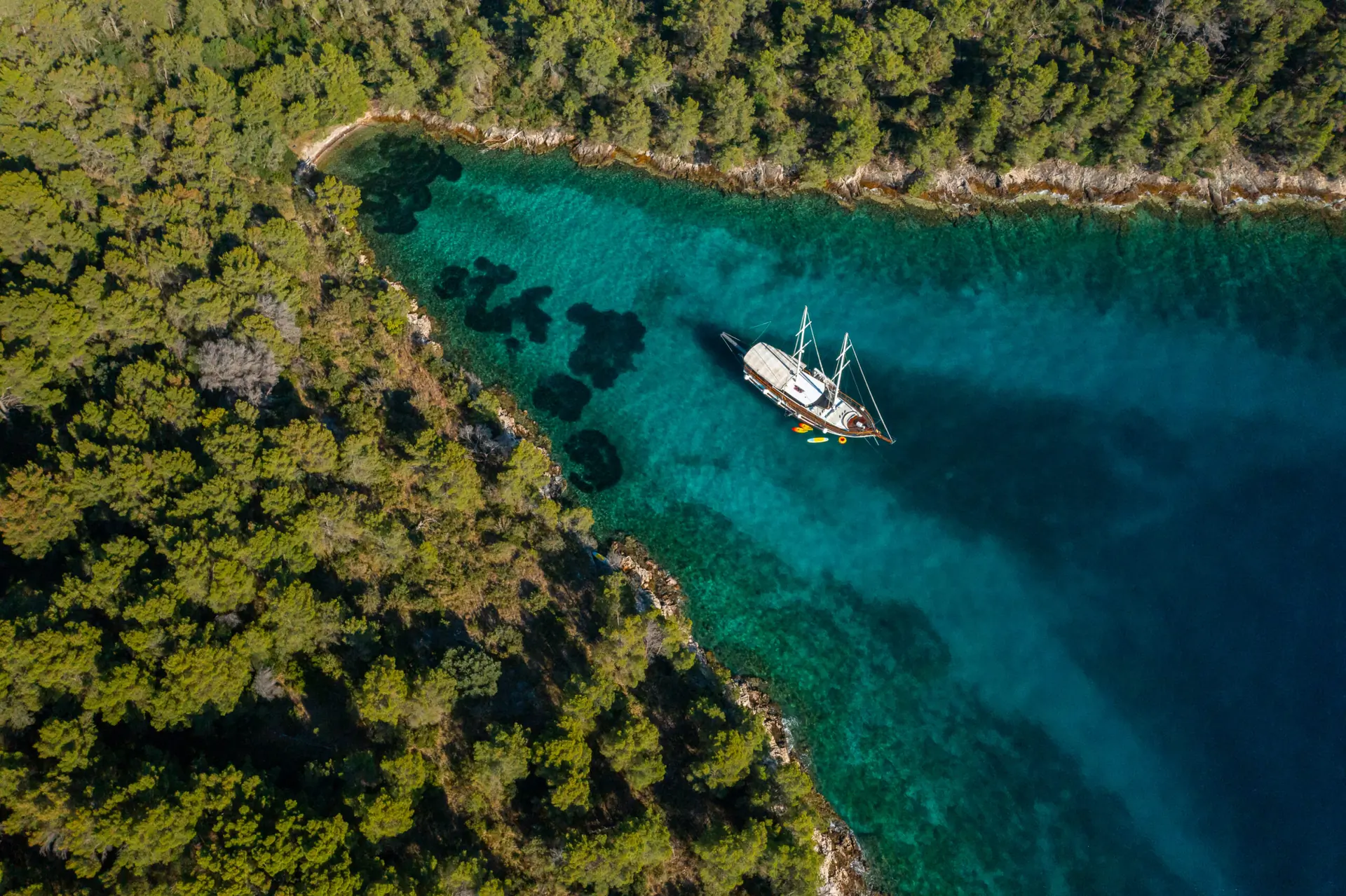 Aerial view of a sailboat anchored in clear turquoise water beside a densely forested shoreline.