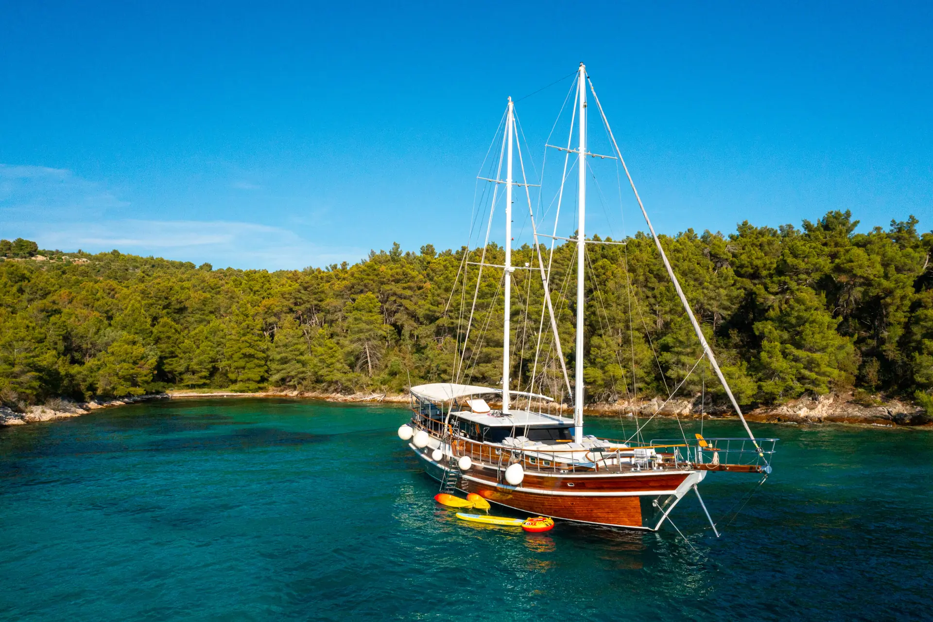 A wooden sailboat with white sails is anchored in clear blue water near a forested shoreline. Paddleboards and a kayak float beside the boat under a sunny, blue sky.