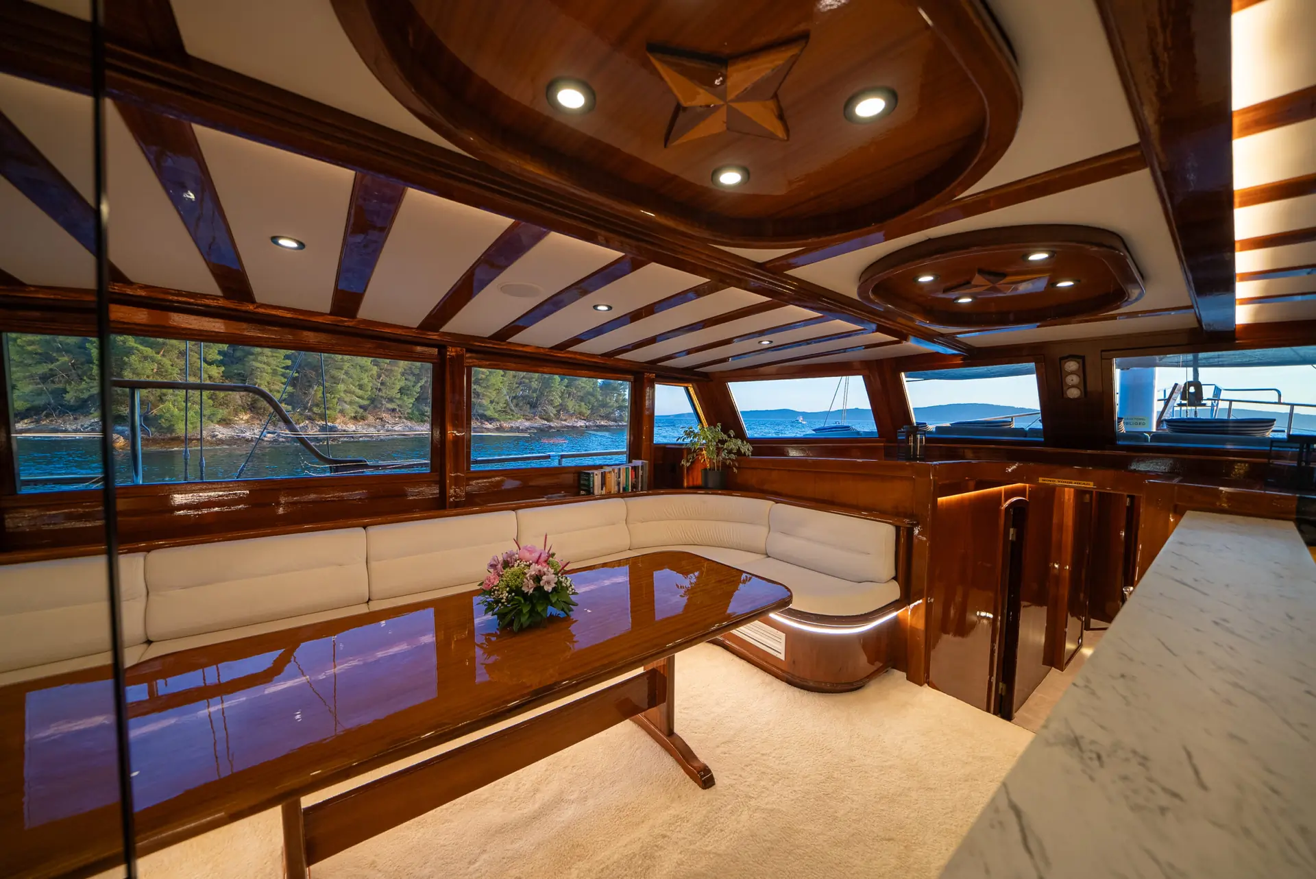 Luxury yacht interior with polished wood, cream-colored seating, a large table, flowers, and large windows offering views of the water and green trees outside. Warm lighting creates an inviting atmosphere.