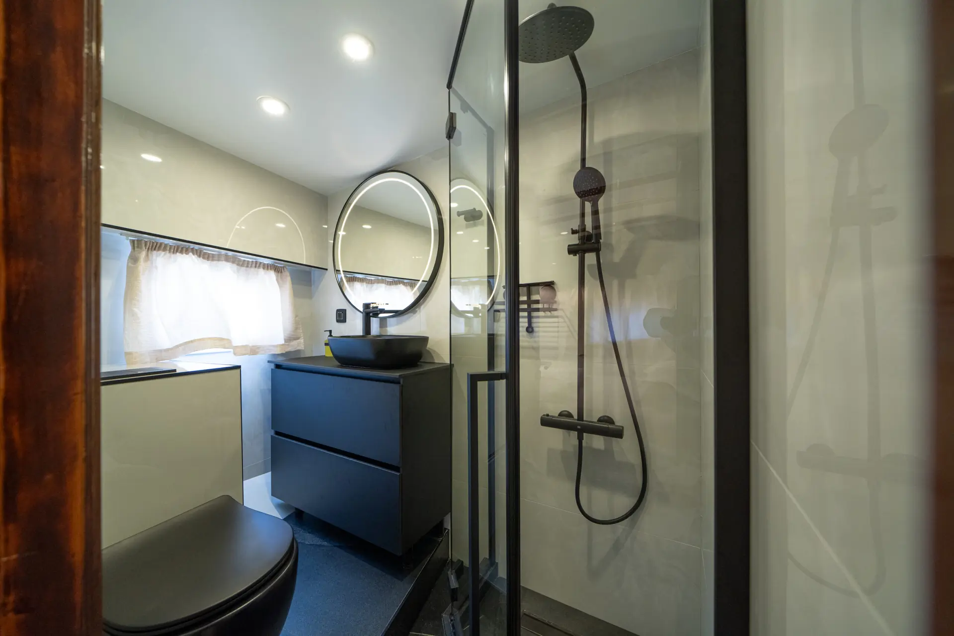 Modern bathroom with a round backlit mirror above a black sink, black drawers, a glass shower enclosure with black fixtures, and a window with white curtains allowing natural light in.