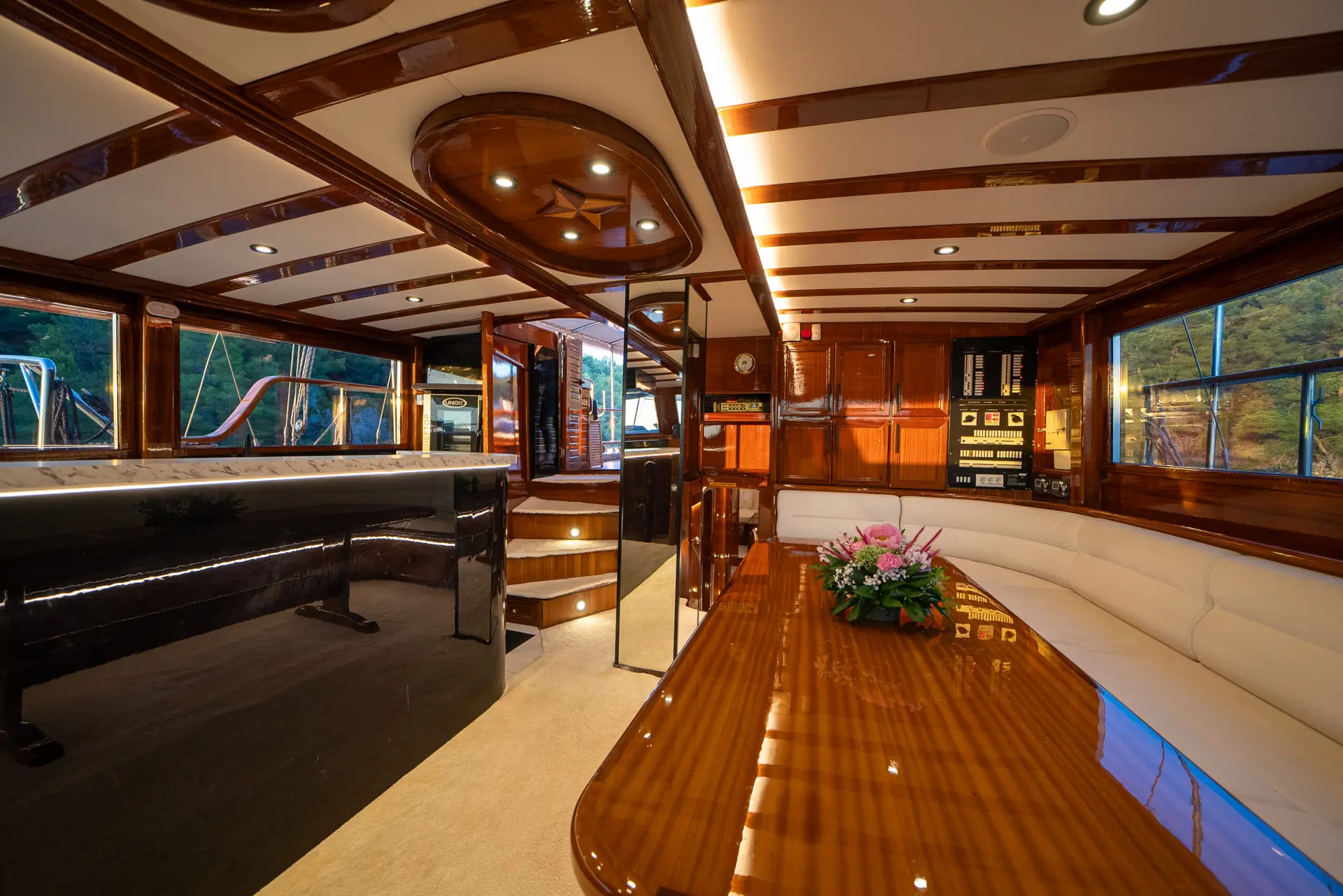 Luxurious yacht interior with polished wood finishes, a long dining table, a curved white sofa, and sleek black countertop. Large windows offer views of water and trees outside. Modern lighting and floral centerpiece decorate the space.
