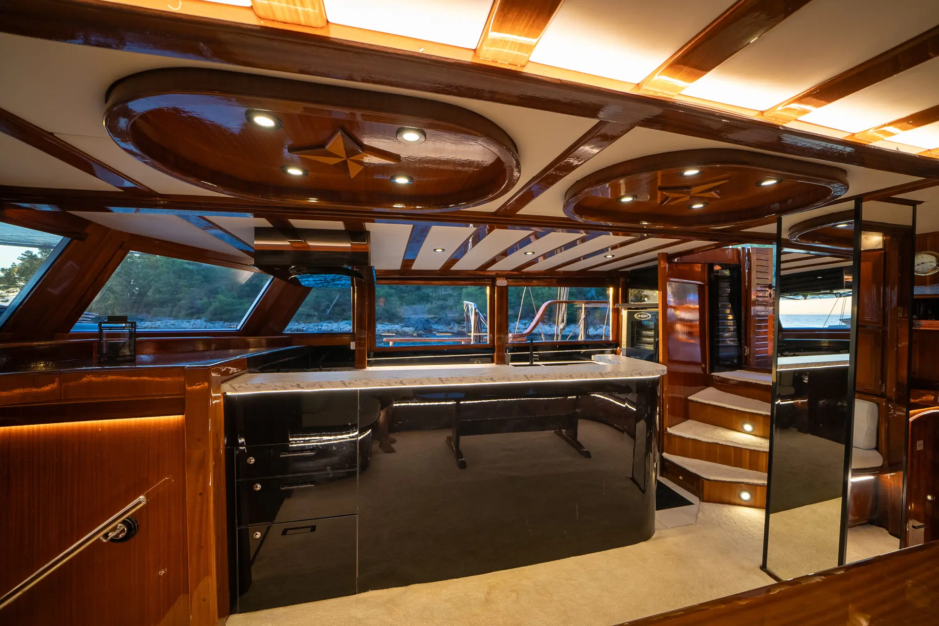 Luxurious yacht interior with polished wood paneling, a sleek black bar, large windows, and carpeted stairs leading to an upper level. Soft lighting highlights the elegant and modern design.