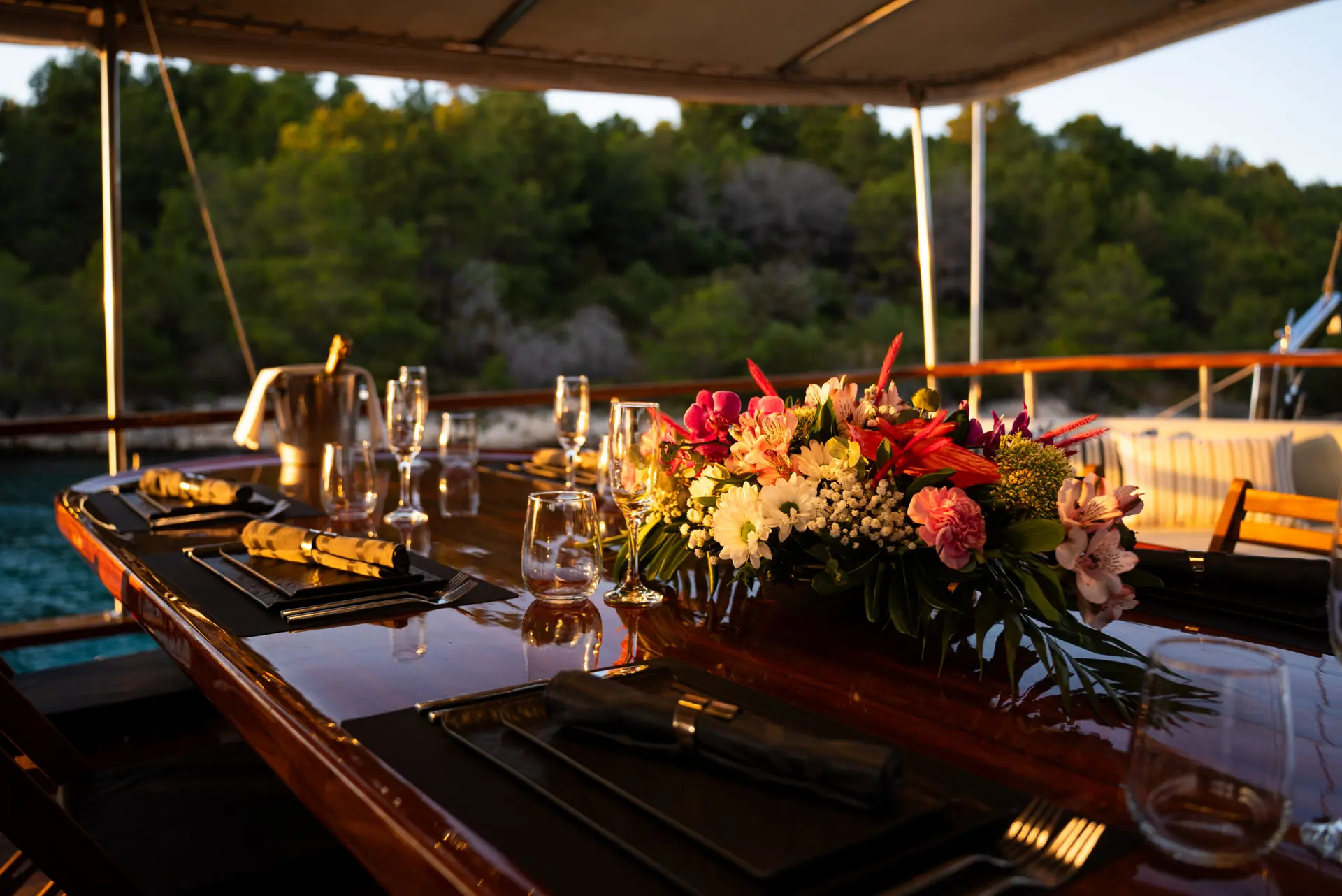 A polished wooden table set for dinner on a boat, decorated with a vibrant floral centerpiece, glassware, and napkins, with trees and water visible in the background.