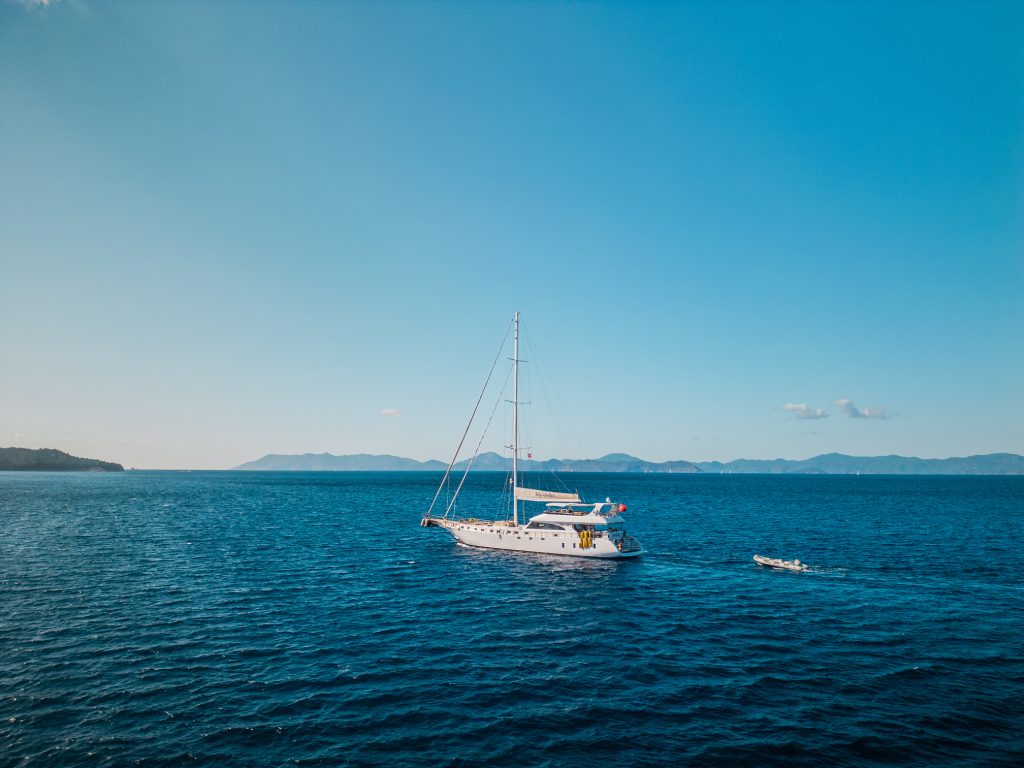 A white sailboat floats on calm blue sea under a clear sky, with distant mountains and a small boat trailing behind.