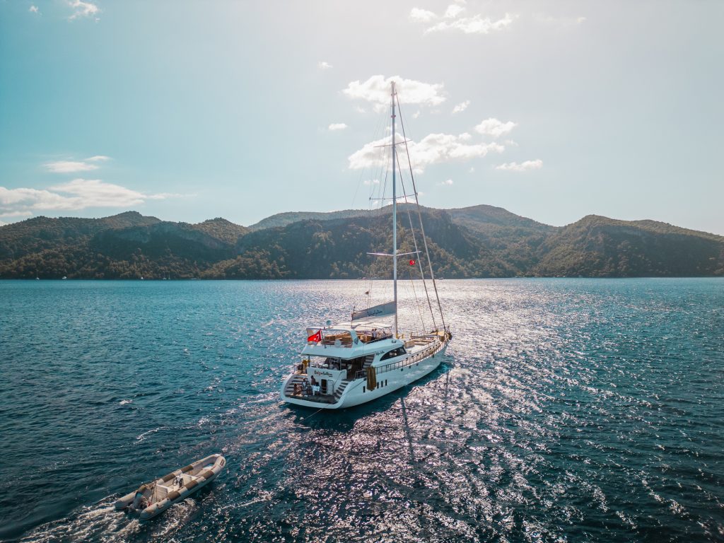 A white sailboat with a Turkish flag sails on a sparkling blue sea, followed by a small dinghy. Forested hills and mountains are visible in the background under a partly cloudy sky.