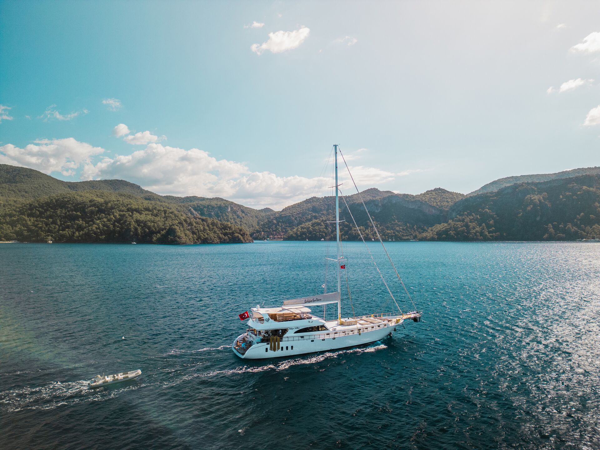 A white sailing yacht cruises on a calm blue sea, surrounded by green, forested hills under a clear sky with scattered clouds. The sun shines brightly, creating a serene and scenic atmosphere.