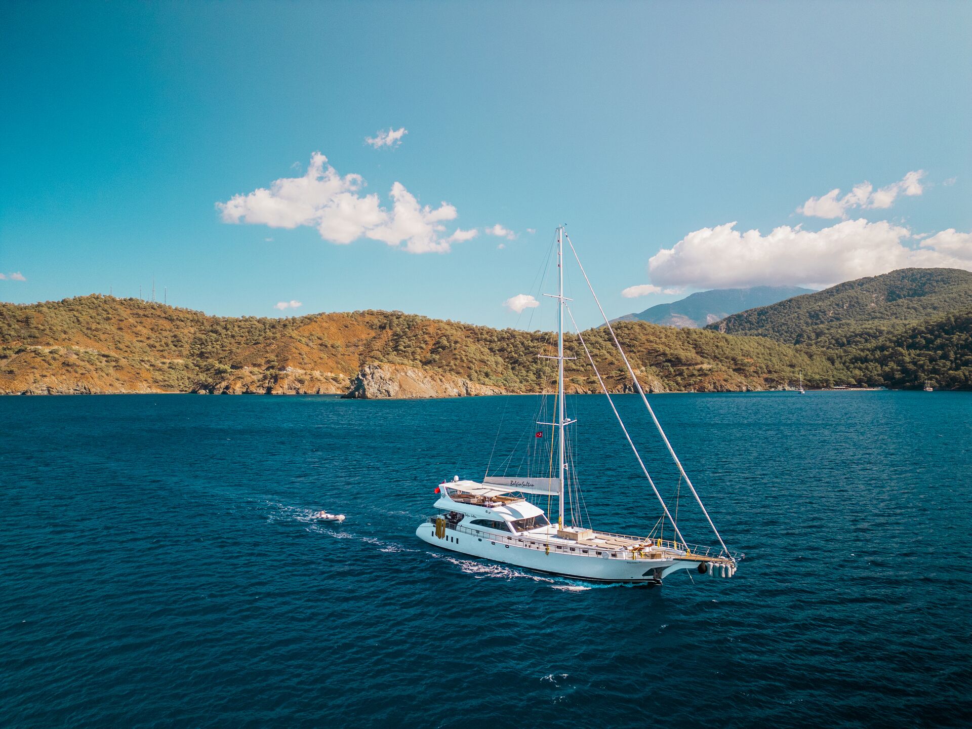 A white sailboat sails on deep blue water near a hilly, green coastline under a clear sky with a few clouds.