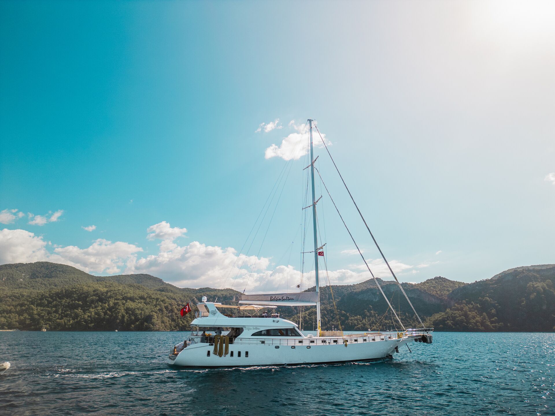 A white sailing yacht with two masts cruises on blue water, surrounded by green hills and under a bright, sunny sky with a few clouds. The Turkish flag is visible at the back of the yacht.