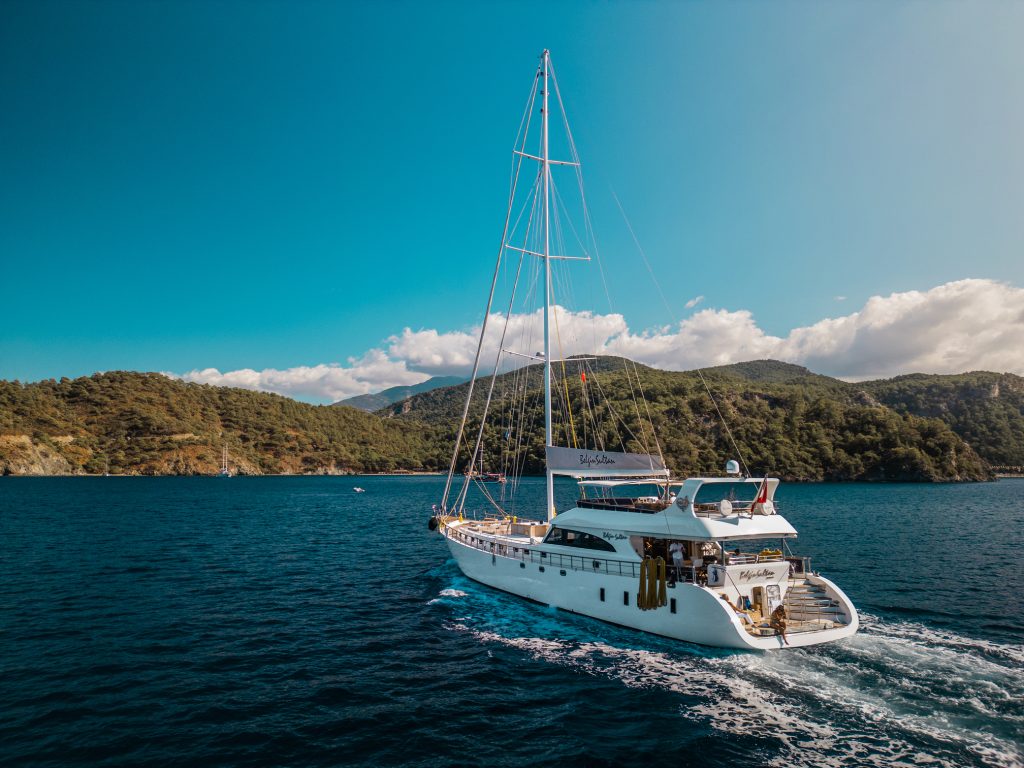 A white sailing yacht cruises on a deep blue sea, surrounded by lush green hills under a clear, bright sky with scattered clouds.
