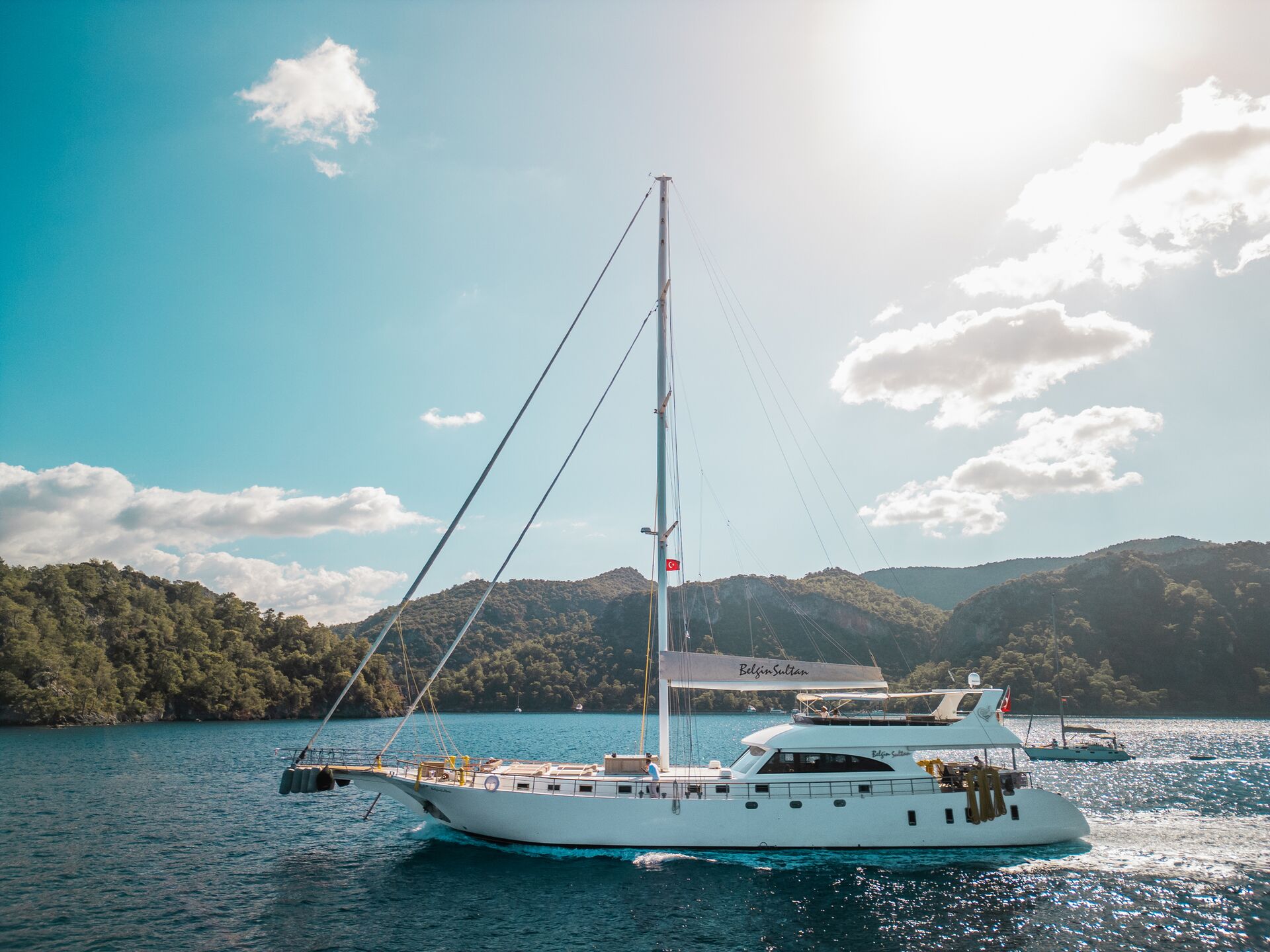 A white sailboat with a canopy floats on clear blue water near forested hills under a bright, sunny sky with scattered clouds.
