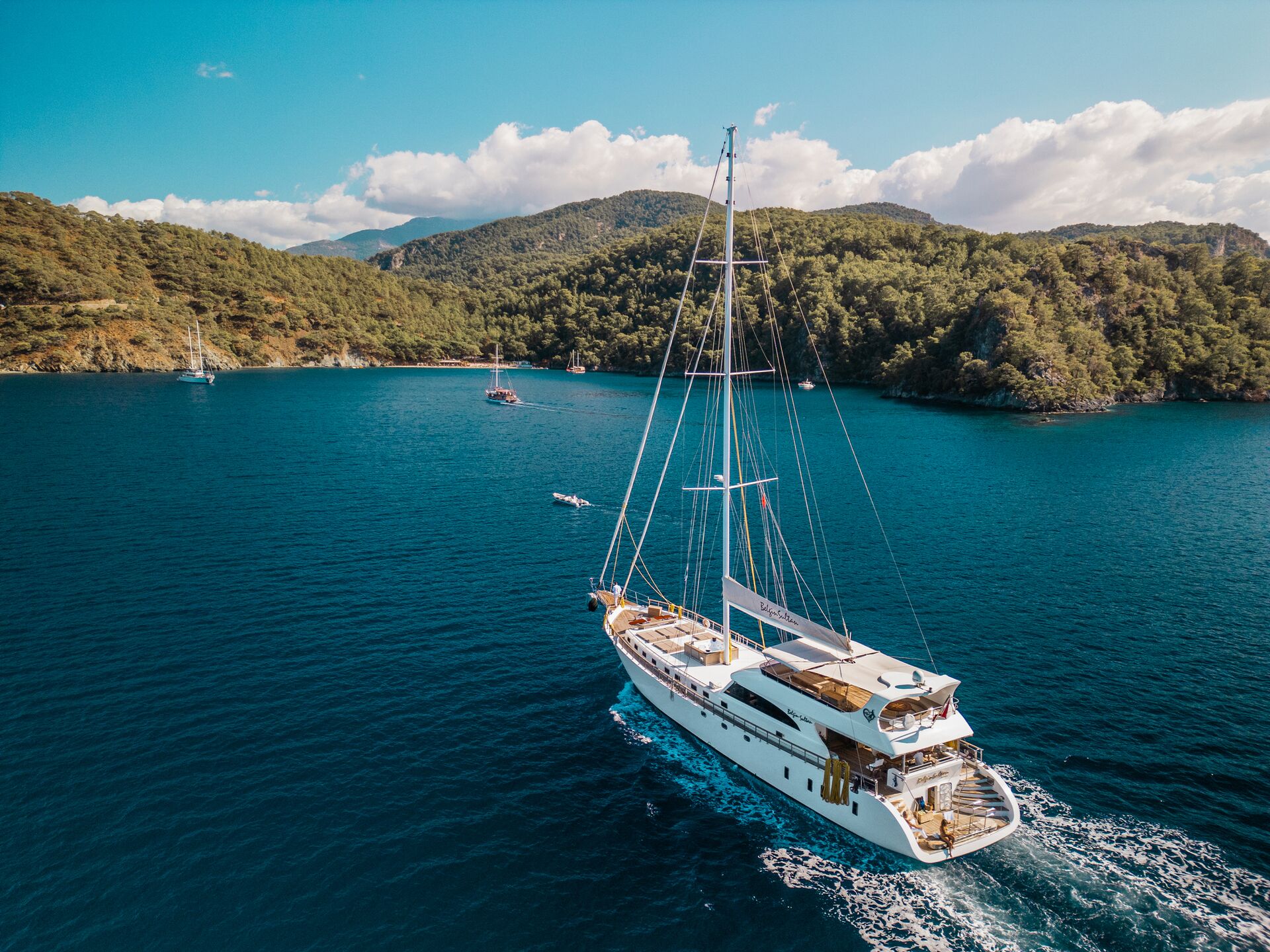 A large white sailboat cruises through calm blue water near a lush, green, hilly coastline under a partly cloudy sky, with other boats visible in the distance.