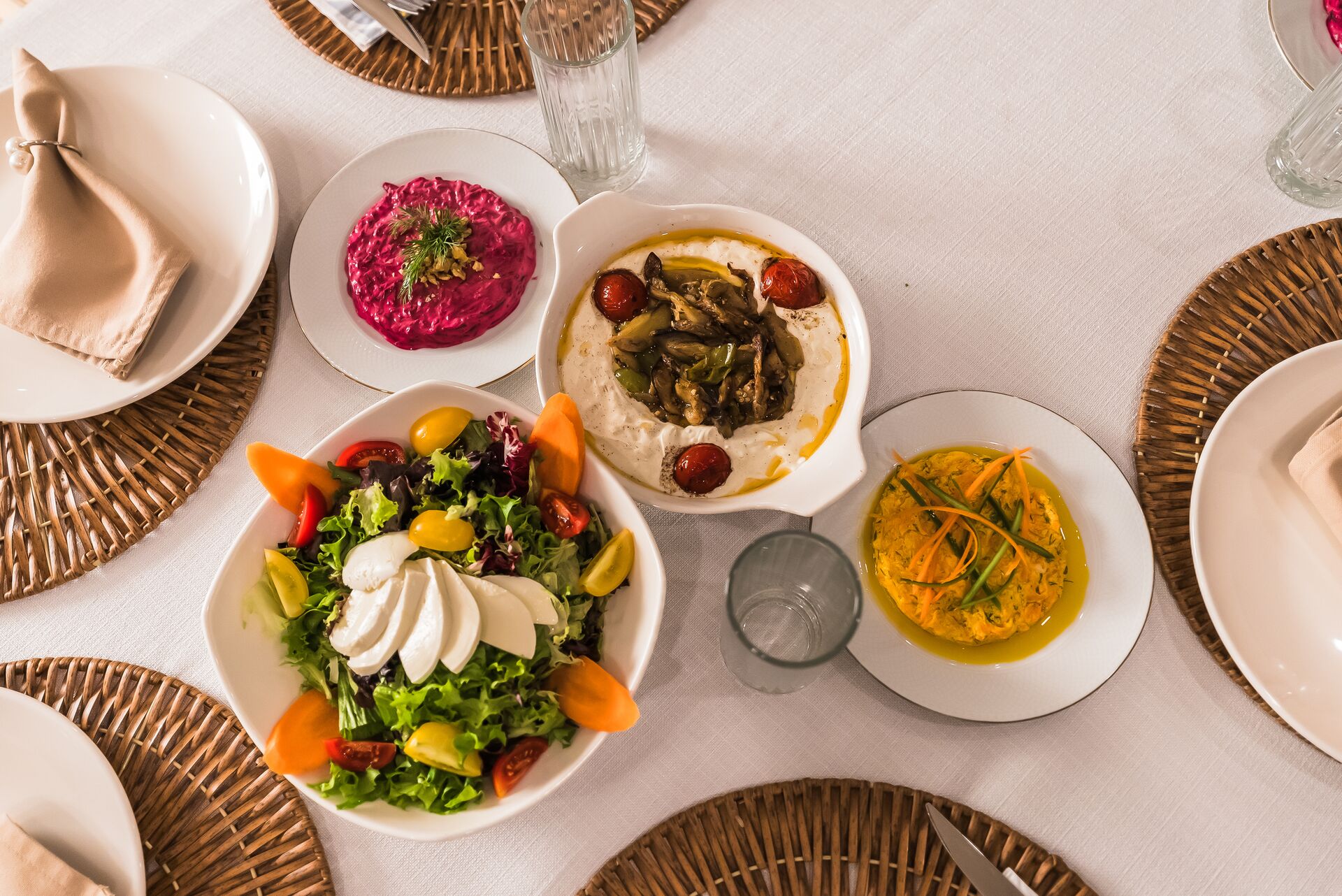 A table set with a fresh salad, a pink beet dip, hummus topped with roasted vegetables and cherry tomatoes, and a yellow carrot salad, all placed on white plates atop woven placemats and a white tablecloth.