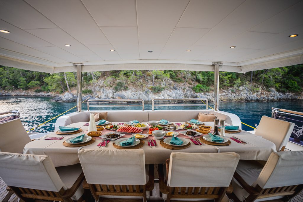 A dining table on a yacht is set with plates, cutlery, and various dishes. The yacht is anchored by a rocky, tree-lined shore, and the seating area is covered by a canopy. The calm water and greenery are visible in the background.