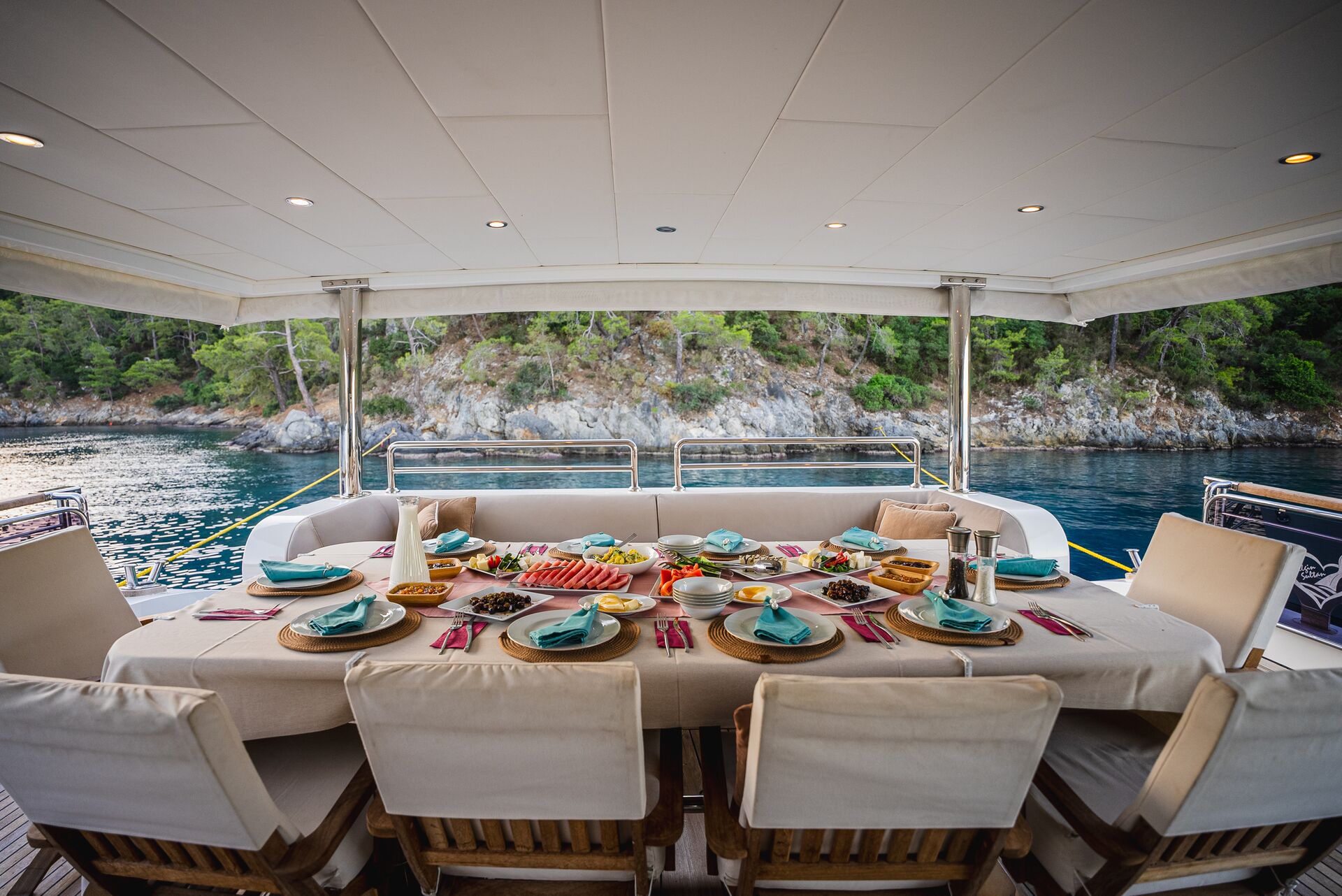 A dining table on a yacht is set with plates, cutlery, and various dishes. The yacht is anchored by a rocky, tree-lined shore, and the seating area is covered by a canopy. The calm water and greenery are visible in the background.