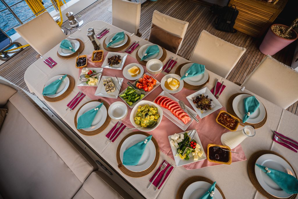Aerial view of a neatly set dining table with plates, cutlery, turquoise napkins, and assorted dishes including sliced fruits, cheeses, vegetables, and sauces, arranged on a boat deck.