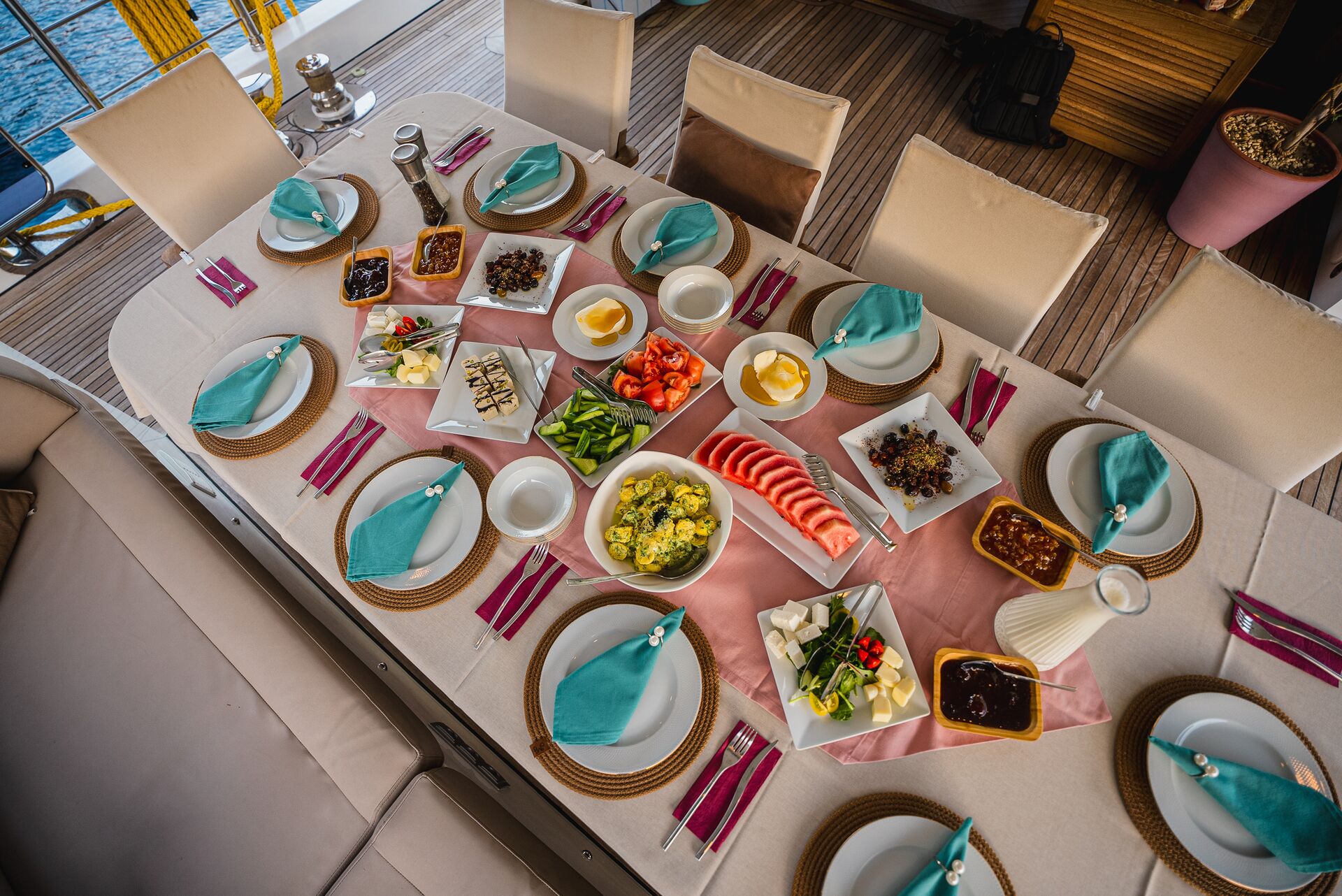 Aerial view of a neatly set dining table with plates, cutlery, turquoise napkins, and assorted dishes including sliced fruits, cheeses, vegetables, and sauces, arranged on a boat deck.