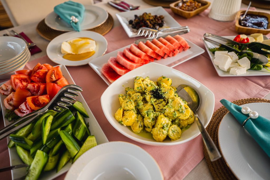 A table set for a meal with plates of sliced cucumbers, tomatoes, potato salad, watermelon, cheese, butter, and small dishes of spreads, on a pink tablecloth with teal napkins.