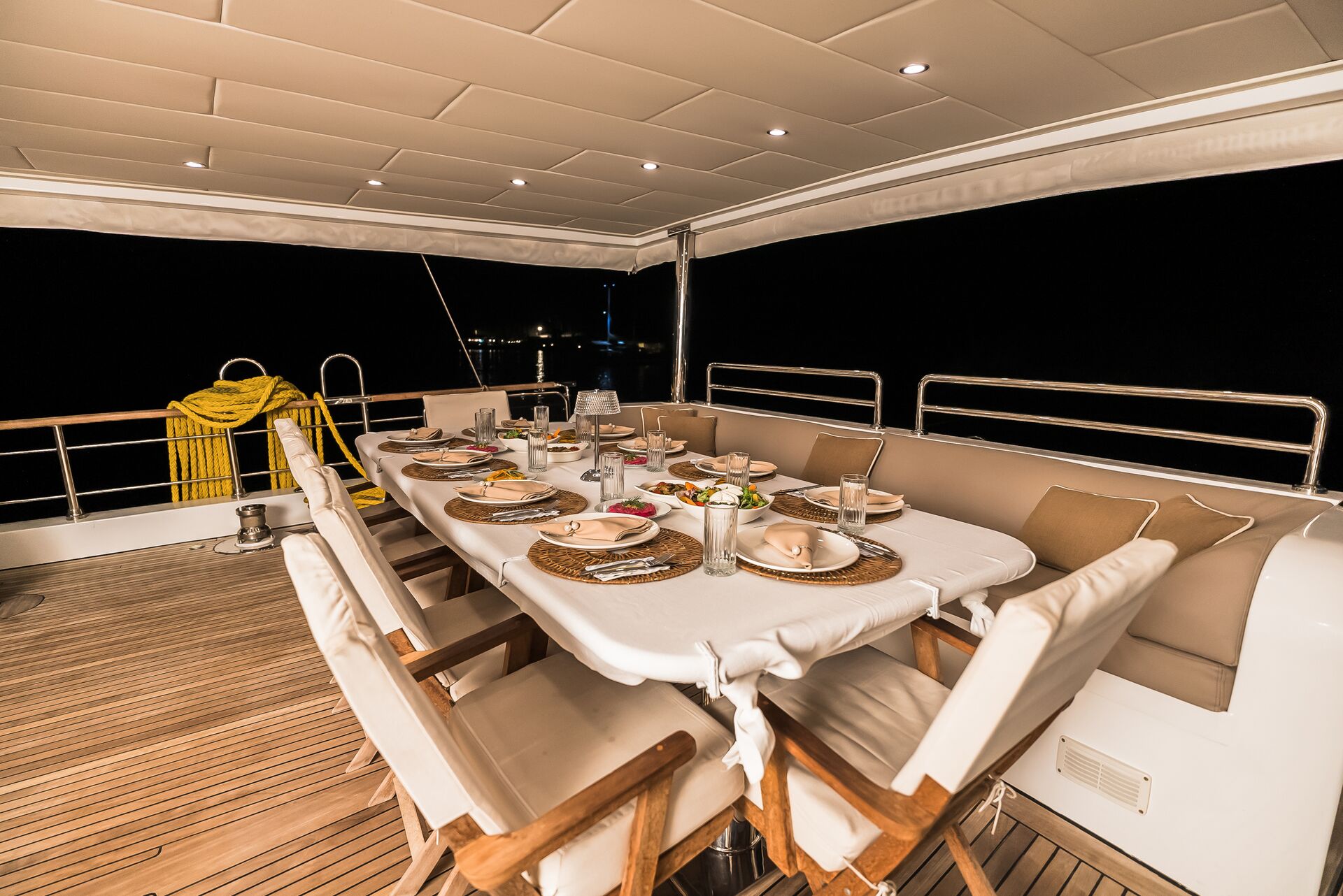 A luxurious yacht deck set for dinner at night, featuring a long table with plates, glasses, and food, surrounded by cushioned chairs. A yellow blanket is draped over the railing, and the background is dark.