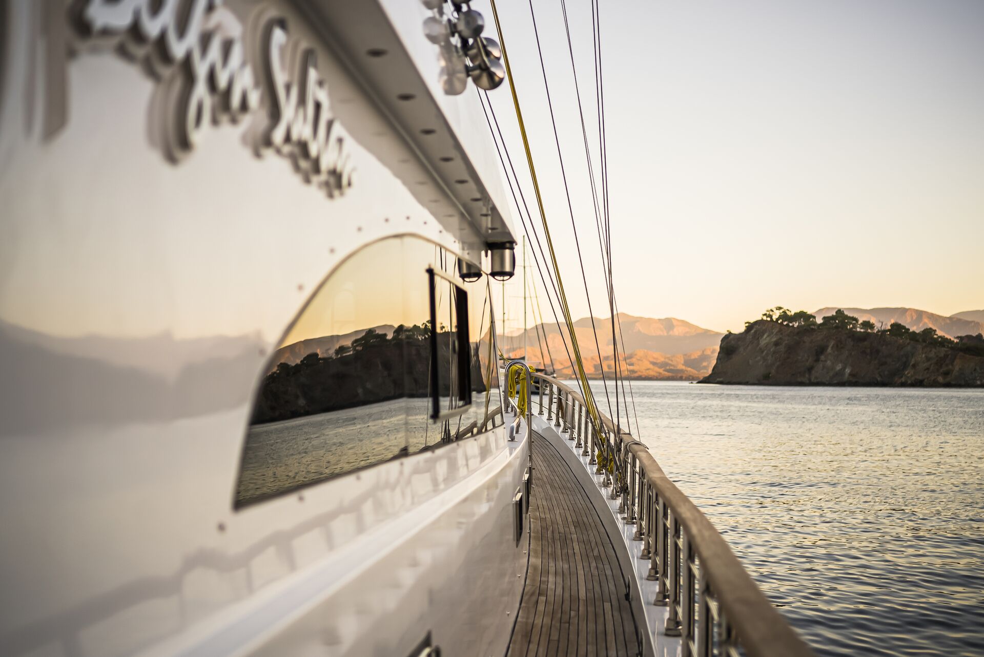 View from the side deck of a yacht sailing near a coastline, with mountains and trees in the distance, calm water, and a warm sunset reflecting on the yachts glossy surface.