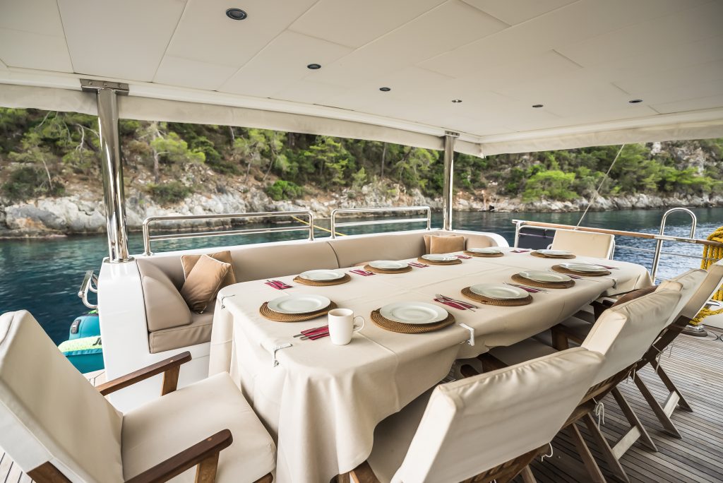 A covered dining area on a yacht with a table set for ten, beige chairs, and a view of calm blue water and a rocky, tree-lined shore in the background.