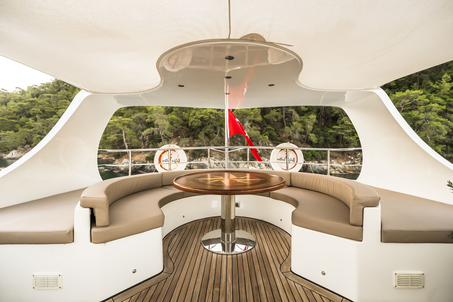 Luxurious yacht deck featuring a round wooden table surrounded by curved beige seating, with a view of the water, green trees, and a red flag at the stern.
